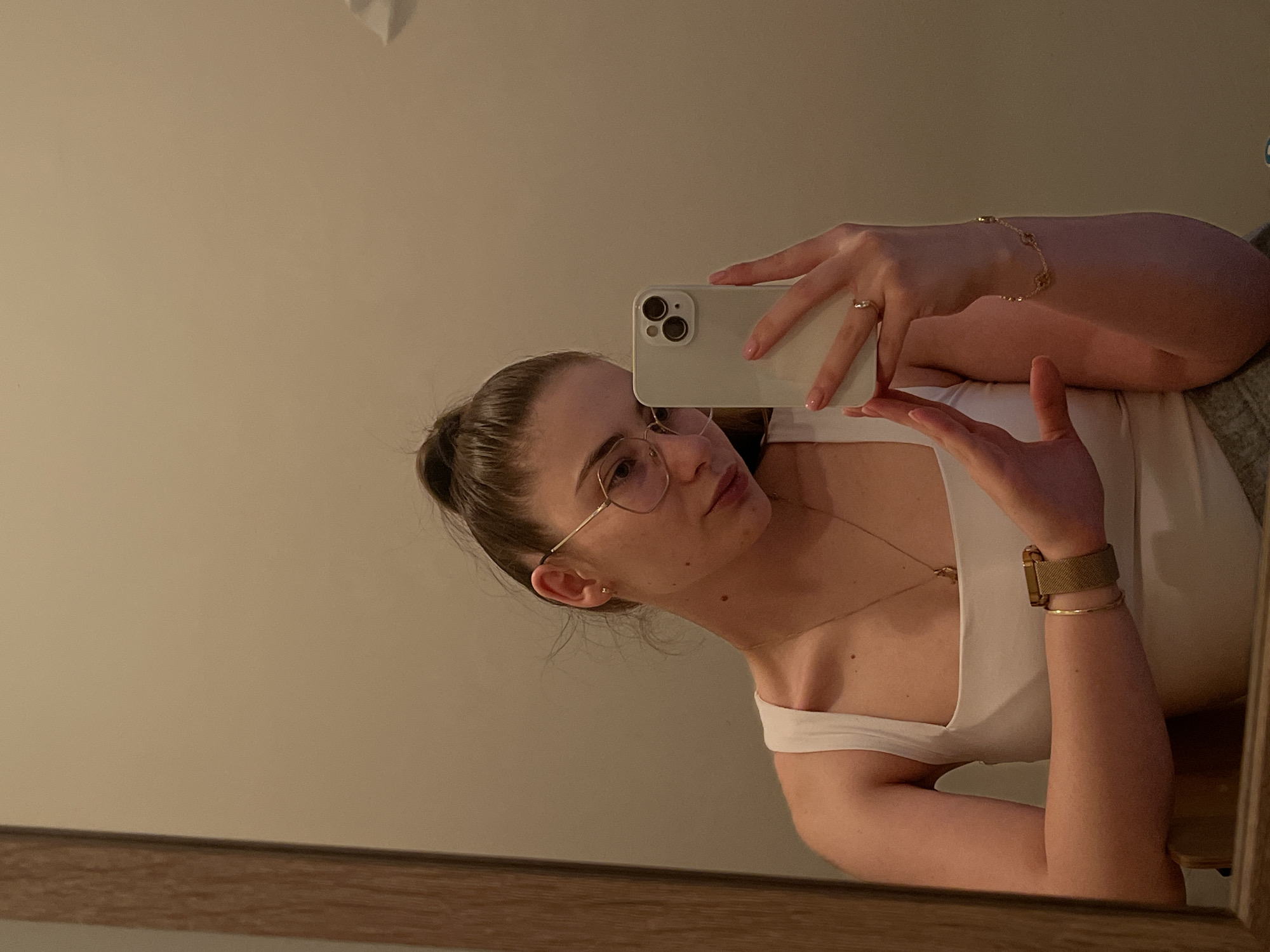 A young woman is captured in a mirror selfie, tilting her head and body to the left. She is indoors, with a plain, light-colored wall serving as the background reflecting in the mirror, and a wooden frame visible at the bottom of the mirror.
The woman is wearing a white tank top or camisole. Her hair is pulled back in a bun, and she is wearing thin, round-framed eyeglasses. Her expression is neutral as she looks towards the phone she is holding. She is holding a light-colored smartphone (likely an iPhone given the camera module design) with her left hand, which has a ring on the ring finger. Her right hand is slightly open with the palm facing up.
Notable accessories include a delicate gold necklace with a small pendant, a thin gold bracelet with small charms on her left wrist, and on her right wrist, a thin gold bangle alongside a wider, gold-toned mesh-style watch band. The lighting is soft and even. No text is visible in the image.