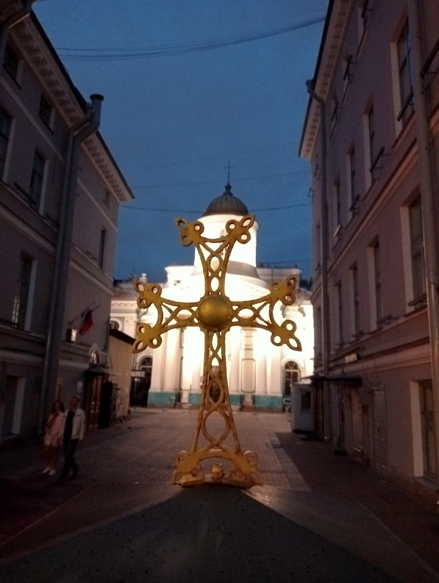 This image depicts a golden cross with ornate decorative elements in the foreground, positioned on a dark, sloped surface. In the background, an illuminated building with a dome and a cross on its roof is visible. The surrounding environment includes buildings on either side of a street or alleyway. Two individuals, a woman and a man, are walking away from the viewer on the left side of the street. The scene appears to be a city street at dusk or twilight. The architecture suggests a location in Saint Petersburg, Russia, potentially near a church or cathedral. The overall ambiance is calm and somewhat serene, with the golden cross acting as a focal point against the subdued lighting of the evening.