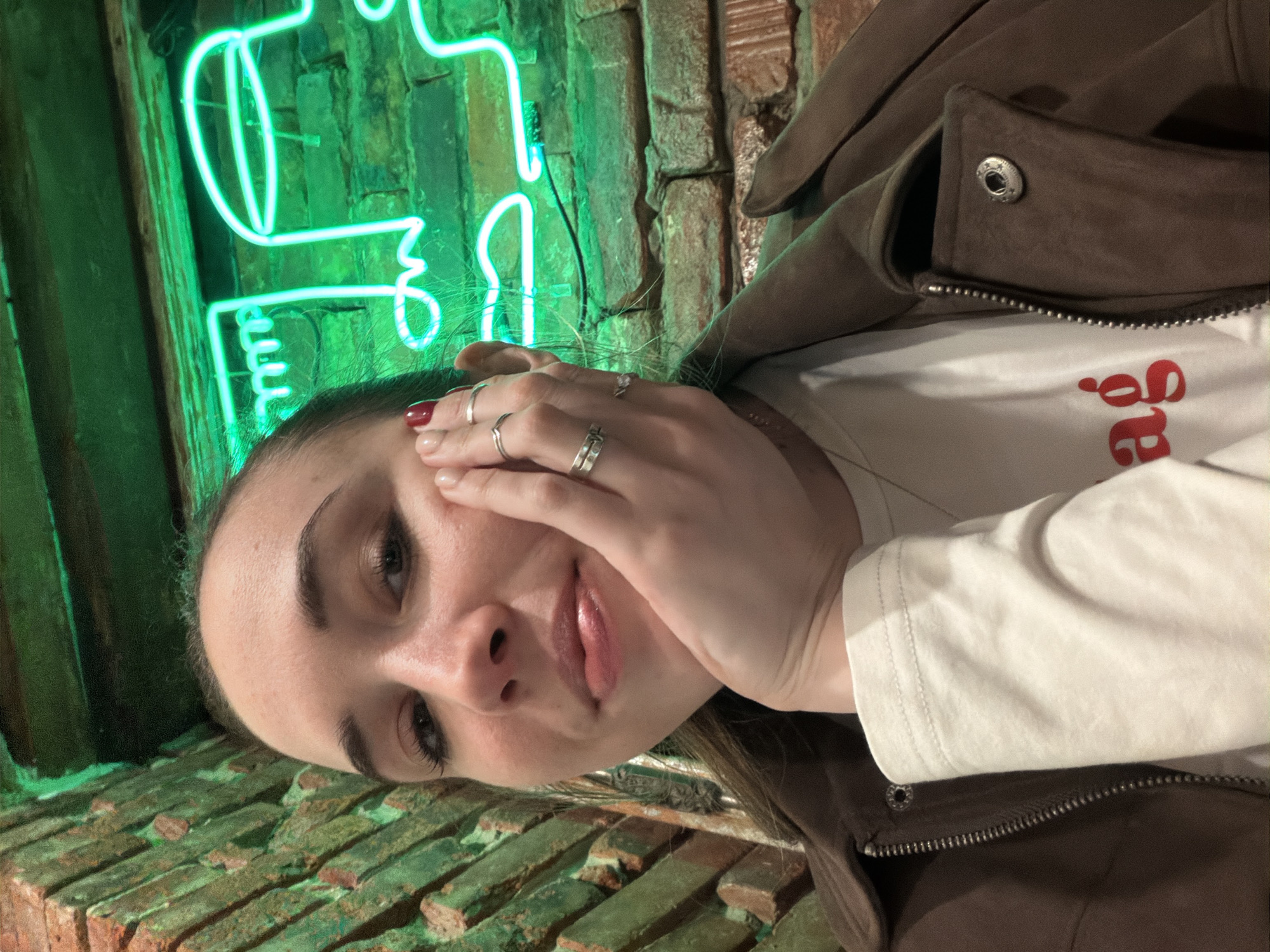 A woman is posing in front of a brick wall with a neon green light in the shape of a stylized face. She is wearing a white long-sleeved shirt under a brown jacket and several rings on her fingers. The background suggests a location in Rostov-on-Don, Russia.