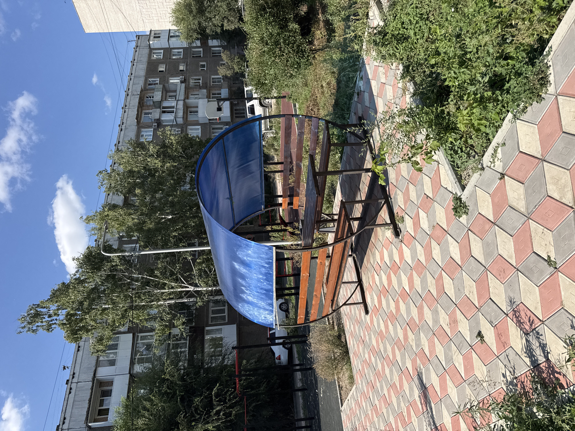 The image shows an outdoor public space in Oskemen, Kazakhstan, with a covered picnic table and benches in the foreground. The picnic area has a curved blue plastic roof and wooden seating. It is situated on a patterned paved area with a geometric design of interlocking diamond-shaped tiles in red, gray, and beige. Behind the picnic area, there are trees and bushes, and in the background, several multi-story apartment buildings are visible. The sky is blue with some white clouds. A white car and a white SUV are parked in the distance. The scene appears to be in a residential courtyard or a small park.
