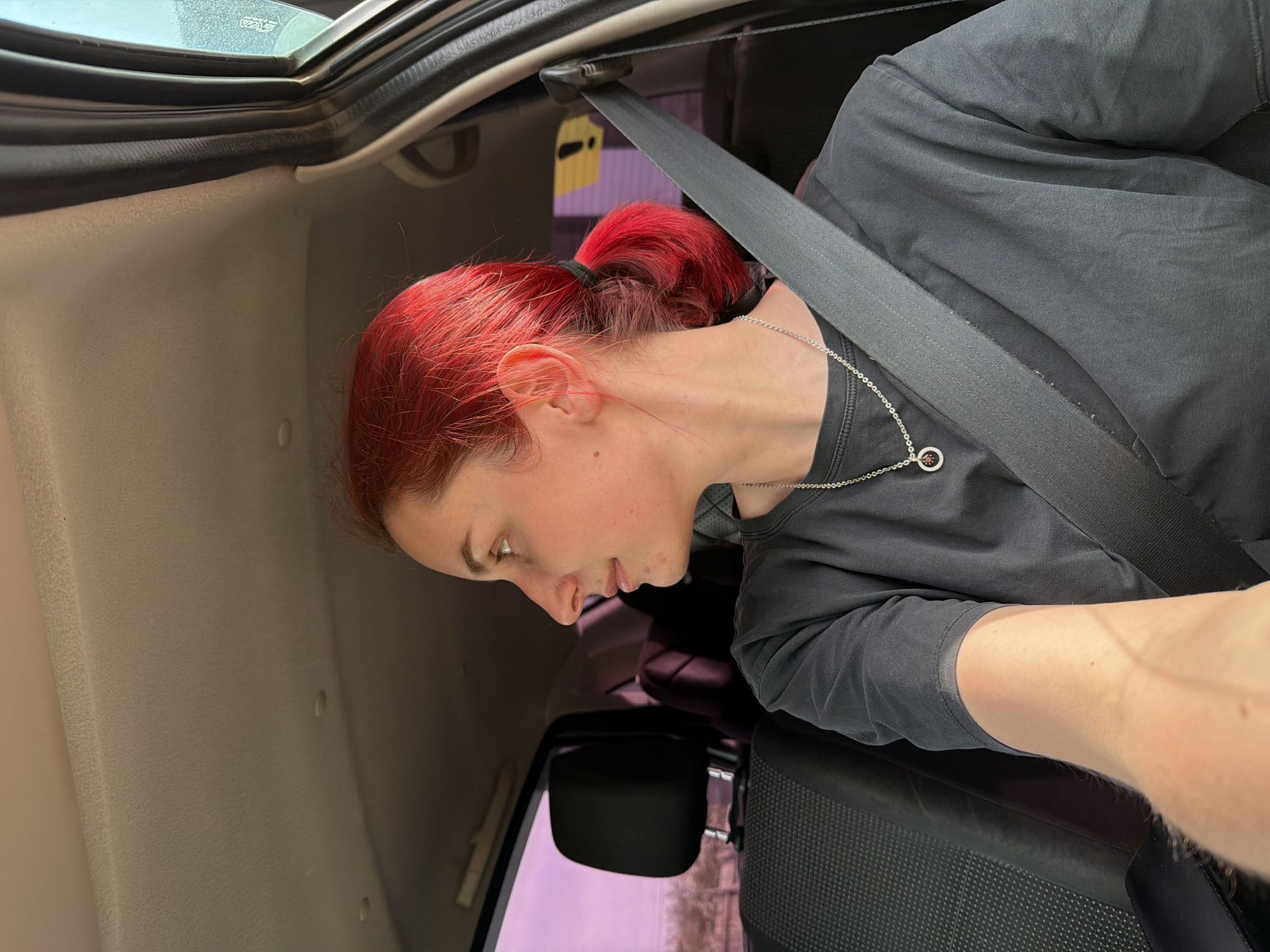 A woman with red hair pulled back into a ponytail is inside a vehicle. She is wearing a dark gray t-shirt, a silver necklace with a circular pendant, and a seatbelt. The interior of the vehicle is light colored, with a black sunroof frame. The woman is looking down towards the left side of the frame. Outside the car, part of a yellow building and a violet-tinted window can be seen. The location cannot be verified as Chaikovsky, Russia, based on the given image.