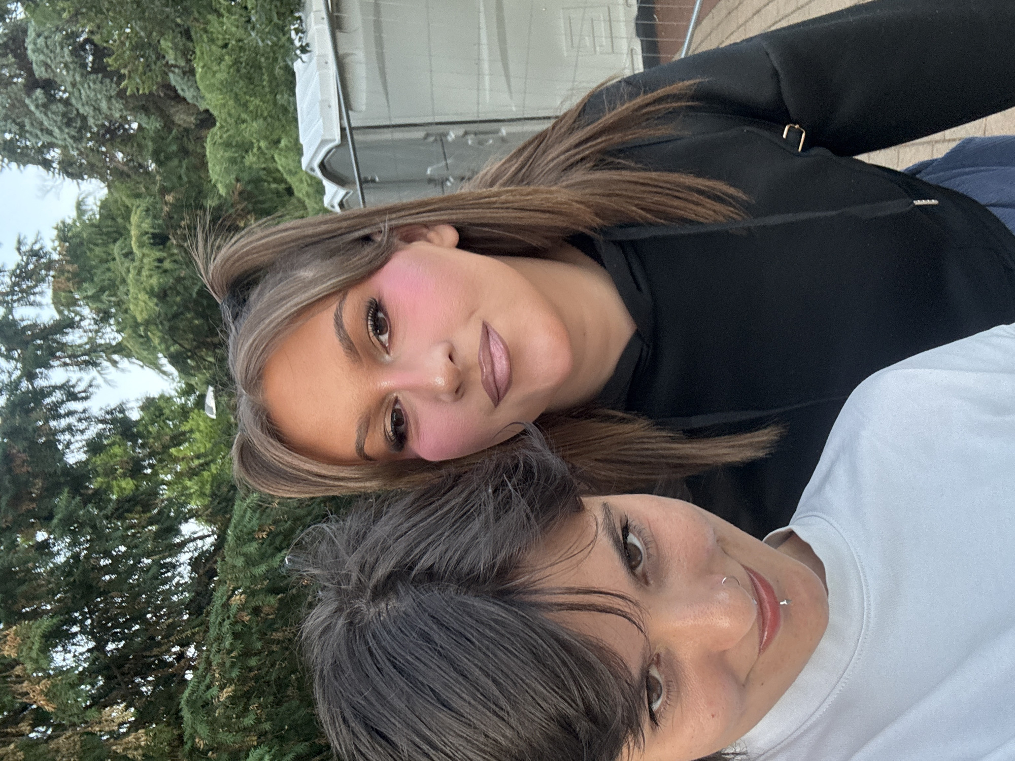 Two individuals are captured in a close-up selfie against an outdoor backdrop. The person in the foreground, on the left, has short, dark, tousled hair and is wearing a white t-shirt. They have a nose ring and a lip piercing. Their gaze is directed towards the camera with a slight smile. To their right is another person with long, light brown hair styled in two high pigtails. They are wearing a black hooded sweatshirt and have prominent pink blush applied to their cheeks. Their eyes are looking forward. In the background, there are trees and what appear to be portable toilets, suggesting an outdoor event or public space. The overall lighting suggests it is daytime. The location is specified as Sofia, Bulgaria.