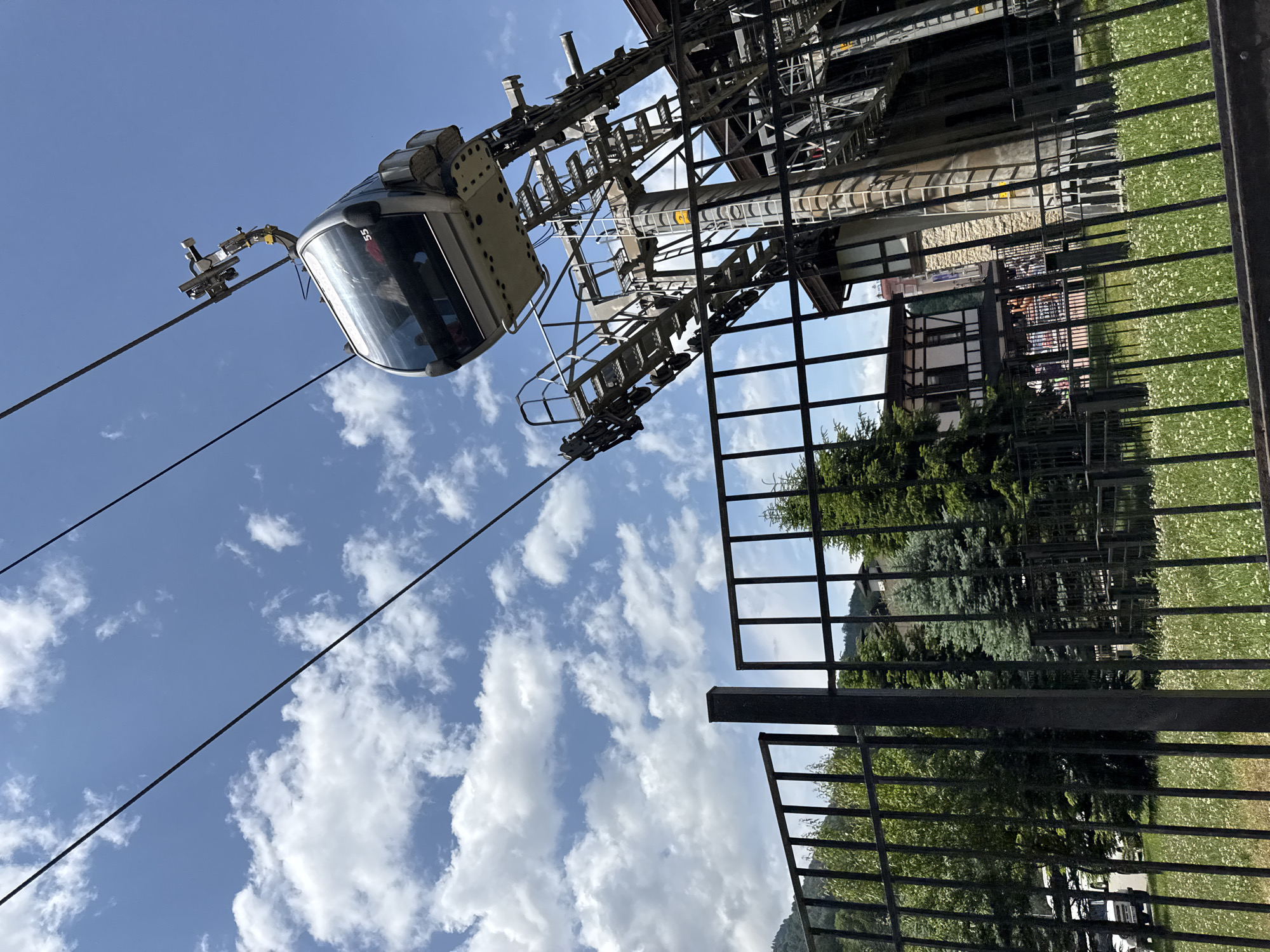 A gondola car labeled "55" is suspended from a cable, moving towards the right of the frame. The gondola is grey with large windows, and several passengers are visible inside. It is attached to a complex mechanical support structure that extends downwards. In the foreground, a black metal fence runs horizontally across the image, with a grassy area dotted with small white flowers behind it. Beyond the fence, trees and a building with a traditional alpine architectural style are visible. The sky is blue with scattered white clouds. The scene is located at a ski resort in Estosadok, Russia, likely near the base station of a ski lift.