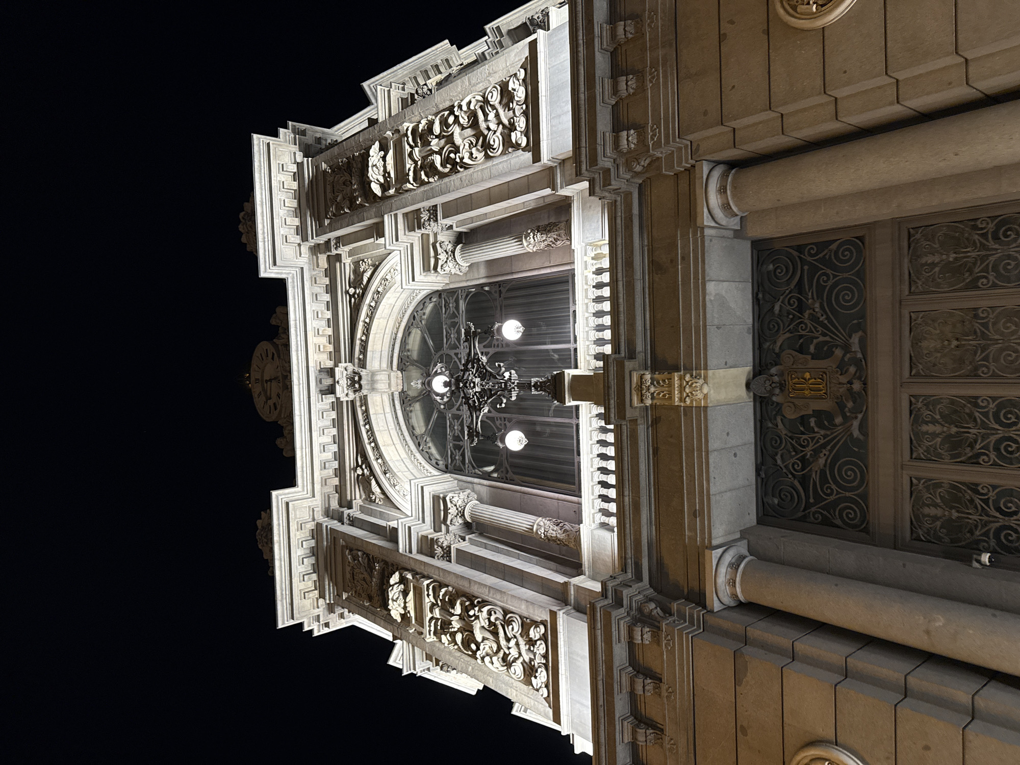 This is a low-angle, nighttime view of the exterior of a grand, ornately decorated building in Madrid, Spain. The building is constructed of light-colored stone and features elaborate architectural details, including carved floral and foliate patterns, columns, and decorative molding. A prominent arched window reveals a chandelier hanging inside. To the right of the window is a large, ornate door with ironwork detailing. A clock is visible on the left side of the building. The background is a dark, clear night sky. The lighting suggests artificial illumination highlighting the building's architectural features.