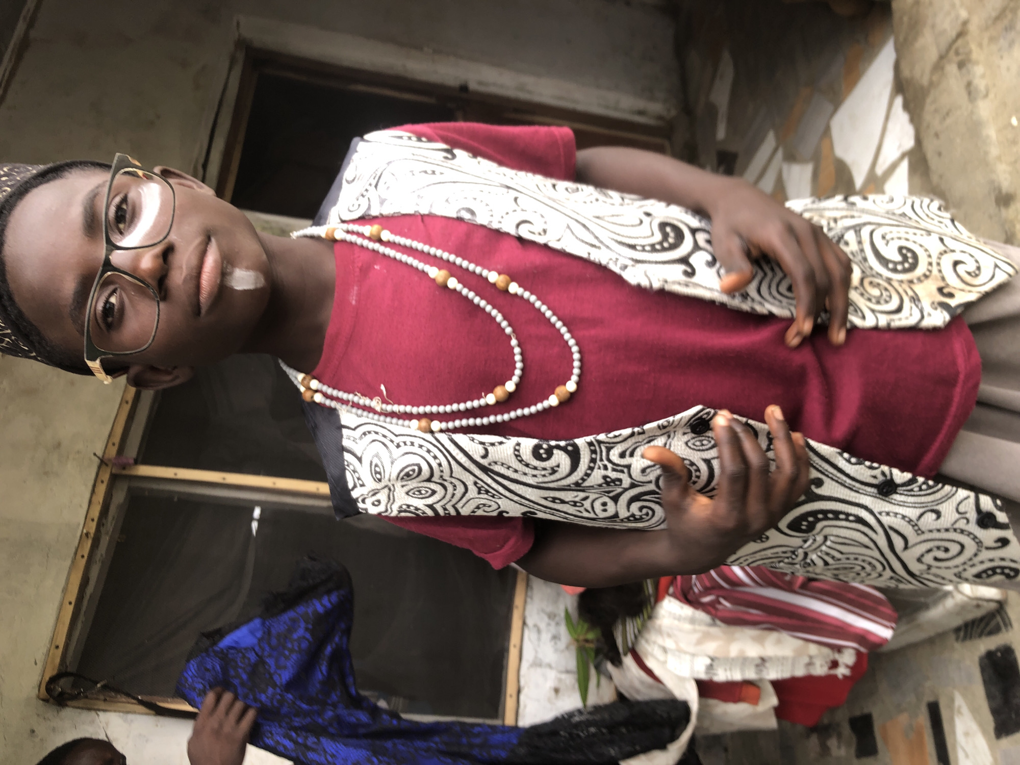 A young Black boy wearing glasses, a red shirt, a black and white patterned vest, and a small cap stands in front of a building. A white stripe is painted on his chin, and he is wearing two beaded necklaces. To the left of the boy, another person is holding up a blue and black patterned garment. In the background, a window with a screen is visible, and beyond that, an exterior door. The ground appears to be paved with tiles. The setting is likely Ado, Nigeria.
