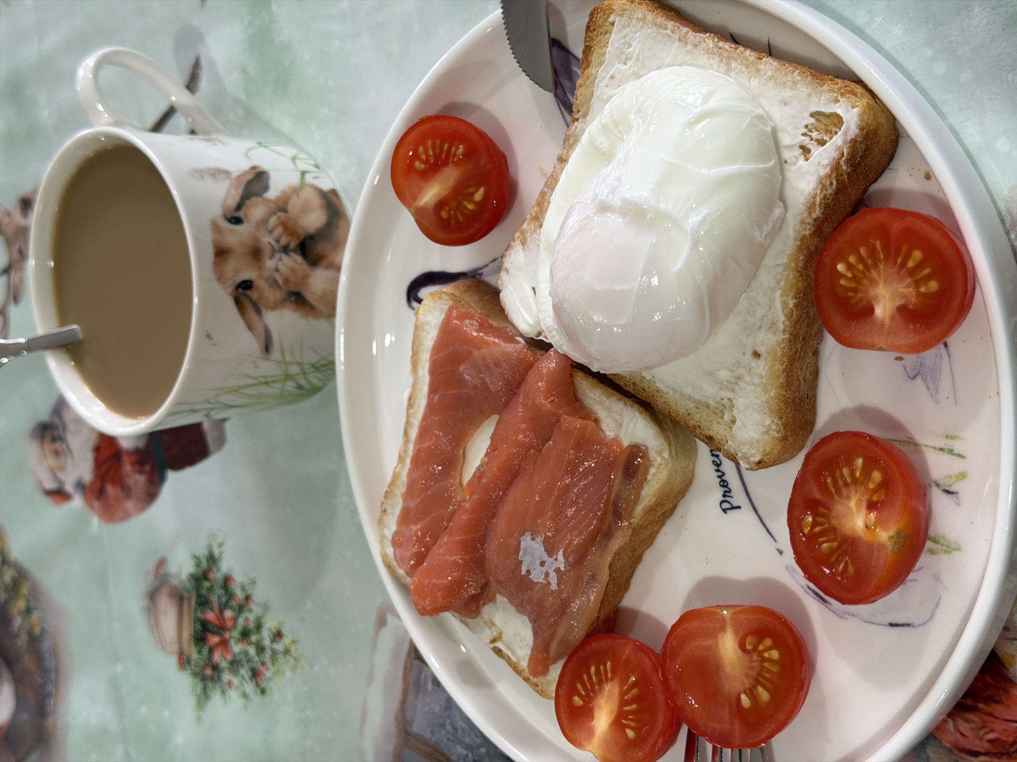 A breakfast scene on a light-colored, patterned surface shows a mug of coffee with a spoon inside, and a plate containing two slices of toast. One slice of toast is topped with a poached egg, and the other is covered with slices of salmon. The plate is also garnished with five tomato halves. A butter knife and a fork are resting on the plate. The mug has a picture of a rabbit on it. The surface beneath the plate and mug has a pattern that includes images of Santa Claus and Christmas trees. The scene is possibly in a home in Chaikovsky, Russia but no specifics are available to confirm its exact location.