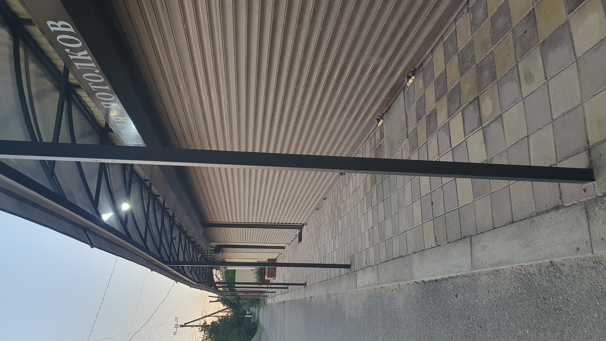 This image depicts the exterior of a building with a covered walkway. The walkway is supported by dark metal beams and has a translucent roof, allowing some light to filter through. Underneath the roof, there are visible light fixtures. The wall of the building behind the walkway is a ribbed, metallic material. The ground in front of the walkway is paved with square tiles in a checkered pattern of grey and beige. A dark, narrow beam runs horizontally across the image in the foreground. The sky is visible in the upper left, suggesting it's either early morning or late evening. In the distance, there are trees and some vegetation, as well as power lines and poles. The overall impression is that of a commercial or public space, possibly an entrance or a shopping area.
There is visible text on a dark sign running along the edge of the covered walkway. The text appears to be in Cyrillic script and reads: "ДЛЯ ПОТОЛКОВ" which translates to "FOR CEILINGS". This suggests the building might be a store or a business specializing in ceiling materials.
The scene is quiet and static, with no people or activity visible. The lighting suggests it is not the middle of the day. The location context is Ust-Labinsk, Russia.