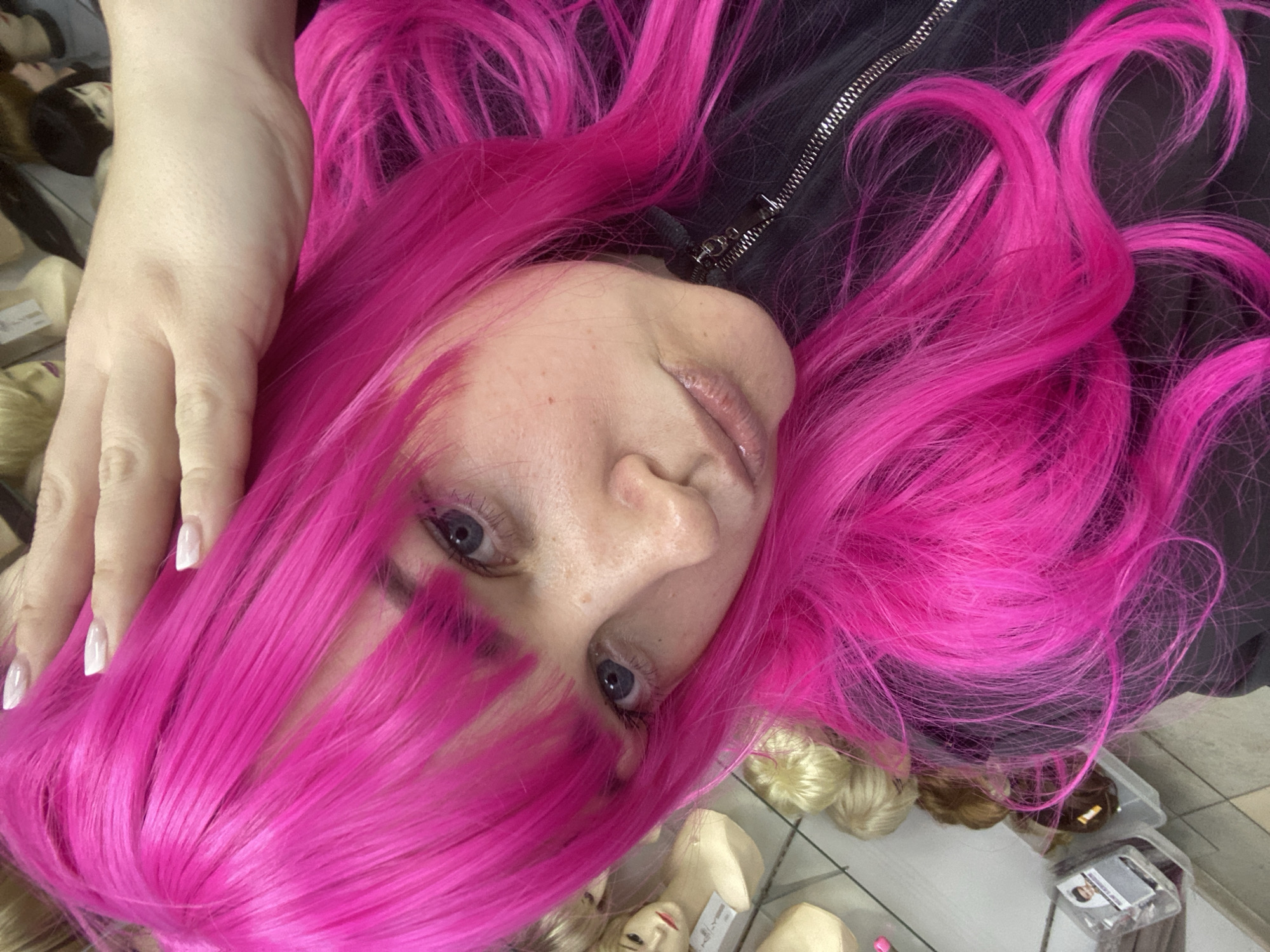 The original image captures a close-up, slightly angled portrait of a person, likely a young adult, with pale skin and light blue eyes, looking directly towards the camera. They are adorned with a strikingly vibrant, long, wavy hot pink wig that features bangs. The person's left hand, with long, white-tipped manicured nails, rests gently on the side of their head, partially nestled within the bright pink hair.
They are wearing a black garment, possibly a jacket or top, with a prominent silver zipper visible on the right side of the frame, near the neck. The background suggests an indoor commercial setting, specifically a wig store or a beauty supply shop, located in Sochi, Russia. This is indicated by several mannequin heads displaying various blonde and brown wigs, as well as loose bundles of hair, scattered across the floor in the lower half of the image. A clear plastic package, possibly for hair products, is also visible in the bottom right corner, though any text on it is illegible. The floor appears to be a light-colored tile. The overall impression is that of someone trying on or showcasing a wig in a retail environment. The time of day is indeterminate, likely indoors with artificial lighting. The person's expression is neutral, consistent with posing for a photograph.