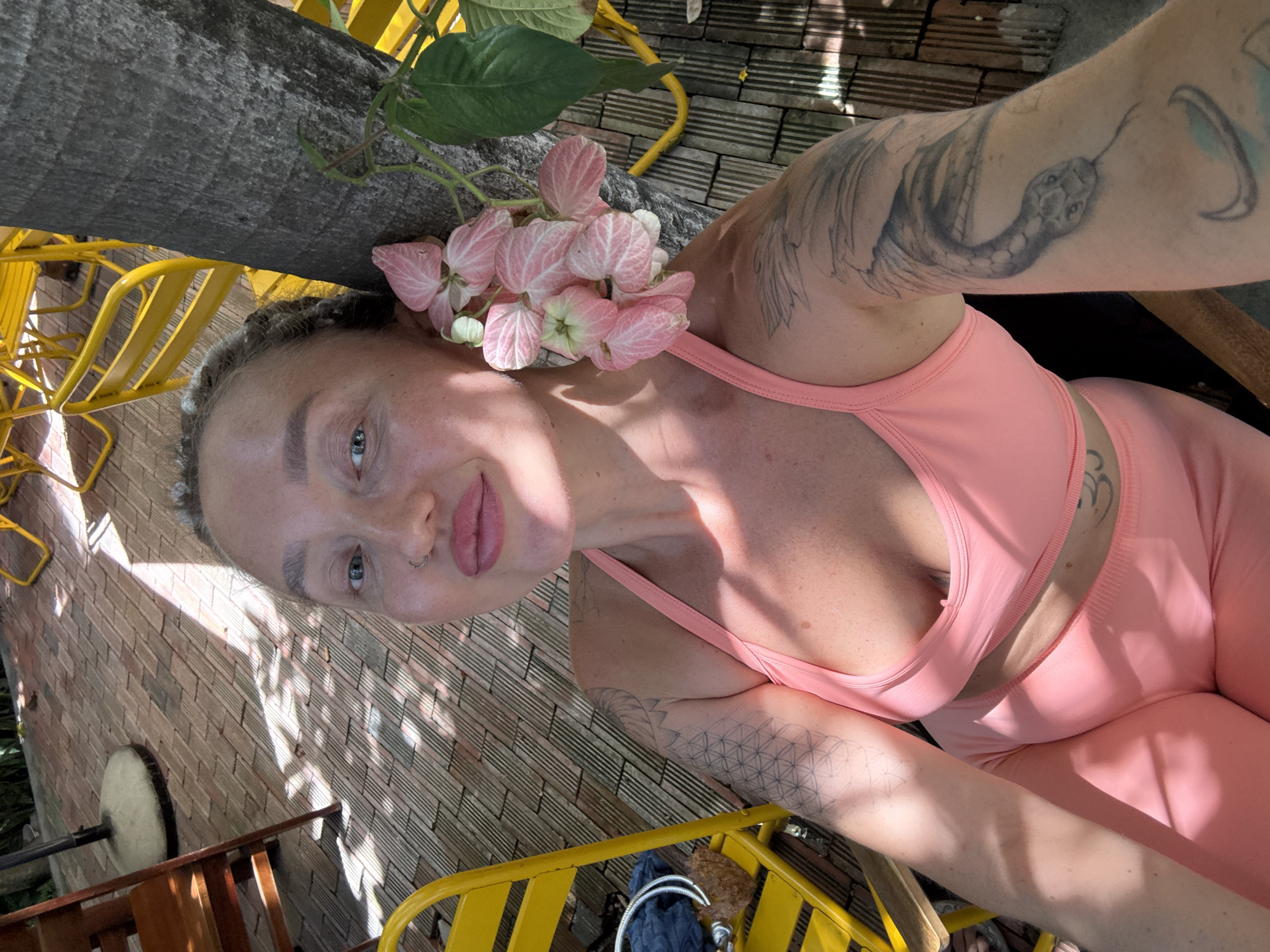 A woman with blonde hair tied back and tattoos on her arms poses for a selfie in a brightly lit outdoor setting. She is wearing a pink sports bra and matching leggings. In the foreground, a cluster of delicate pink flowers with white accents adorns the side of her head, appearing to be tucked into her hair. Her gaze is directed at the camera, and she has a subtle smile. The background reveals a brick wall and a wooden deck, with yellow chairs and a glimpse of greenery from a tree. Sunlight casts dappled shadows across the scene, suggesting it is daytime. The image appears to be taken in Nha Trang Ward, Vietnam.