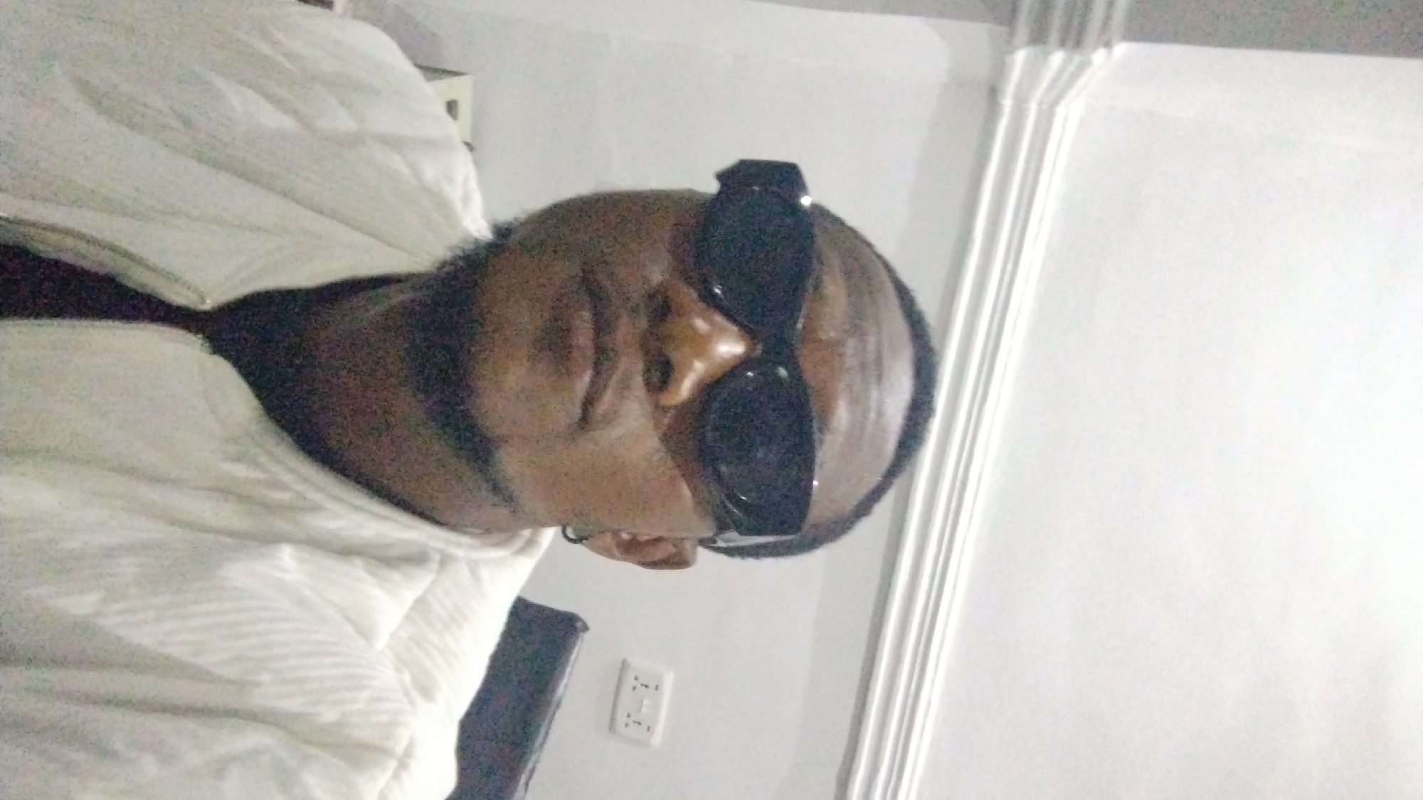 A person wearing sunglasses is reclining in bed, facing the camera. A power outlet is visible on the wall behind the head of the bed. The scene is located indoors, in an unknown location within the city of Unknown, Unknown.
