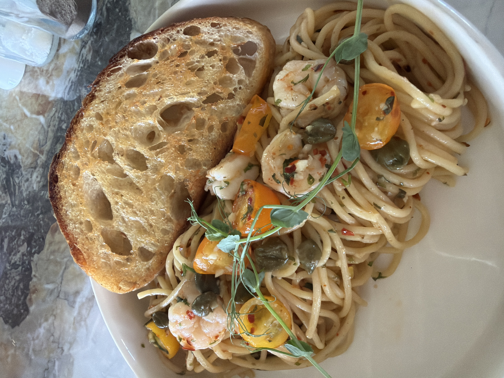 The image depicts a plate of pasta with shrimp, tomatoes, capers, and sprouts, accompanied by a slice of toasted bread. The plate is placed on a marble-like surface, with a glass container possibly holding salt or pepper visible in the background. No people are visible. The location, Kuta Selatan, Indonesia, is not directly discernible from the visual elements present.