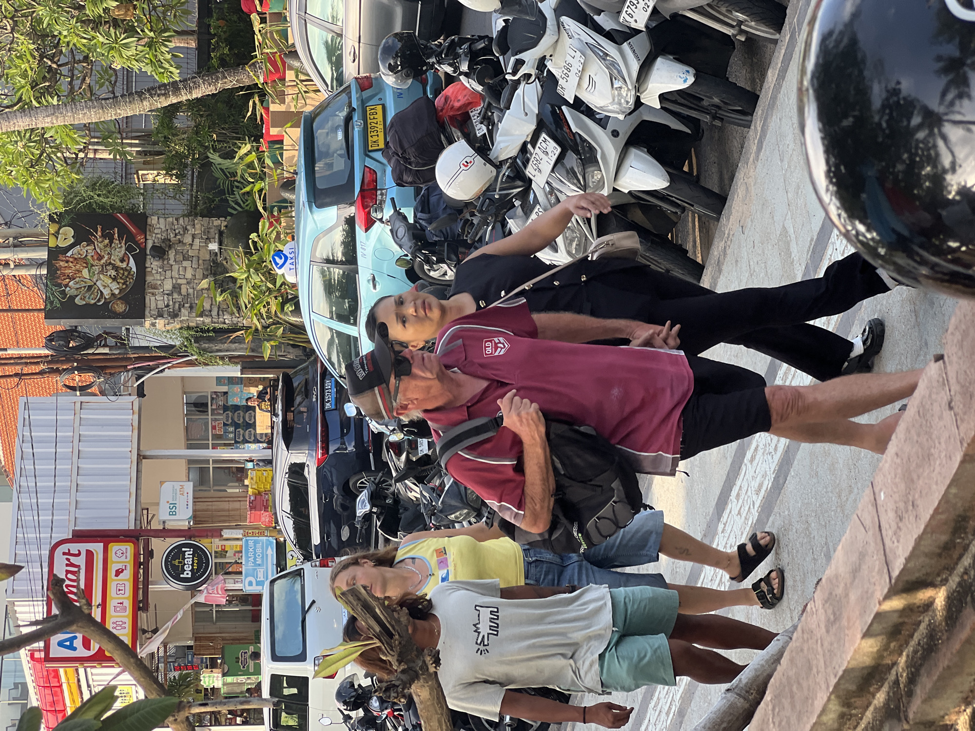 The image depicts a street scene in Kuta, Indonesia, with several people walking along a sidewalk. In the foreground, a person in a white t-shirt and light blue shorts walks towards the left, partially obscured by a piece of wood. Behind them, a woman in a yellow tank top and denim shorts is walking, followed by a man wearing a maroon polo shirt and dark shorts, carrying a backpack. To their right, a woman in a black sleeveless top and black pants is walking.
The background shows a commercial area with buildings housing shops. A prominent sign for "Alfamart" is visible, as well as signs for "BSI" and "bean! SPOT". Several motorcycles are parked along the street, and a blue taxi with the word "TAKSI" on its roof is also present. A grey car is parked to the far right. The environment appears to be an urban street with palm trees visible, suggesting a tropical location. The lighting suggests it is daytime, possibly late afternoon, given the warm tones.