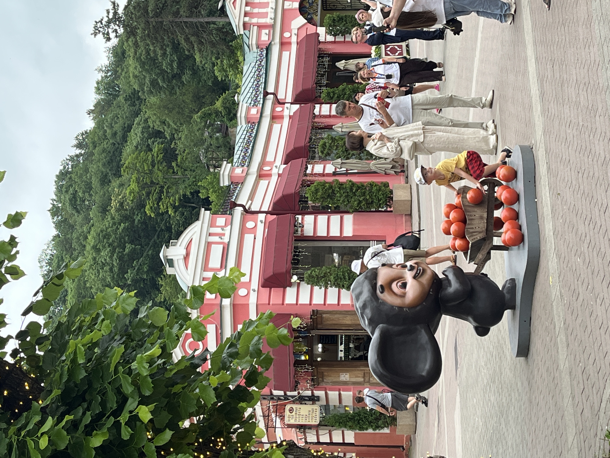 This image captures a lively scene in the city of Kislovodsk, Russia. In the foreground, a large, dark statue of Cheburashka, a popular Russian cartoon character, stands with its mouth open and a playful expression. The statue is accompanied by a wooden cart filled with red, round objects, likely representing tomatoes. A young boy in a yellow shirt and plaid shorts is positioned near the cart, reaching for one of the objects.
Behind the statue, a crowd of people mills about. Many are casually dressed, suggesting a pleasant day outdoors. A striking pink building with white trim and red awnings dominates the background. The building appears to be a shop or restaurant, with an open doorway visible. Green trees and a hill covered in lush foliage rise behind the structure, indicating the natural beauty of Kislovodsk. The paving stones of the walkway are visible in the foreground, creating a textured ground. The overall atmosphere is one of a public gathering, possibly in a park or entertainment area, with elements of both cultural interest (the statue) and everyday life.