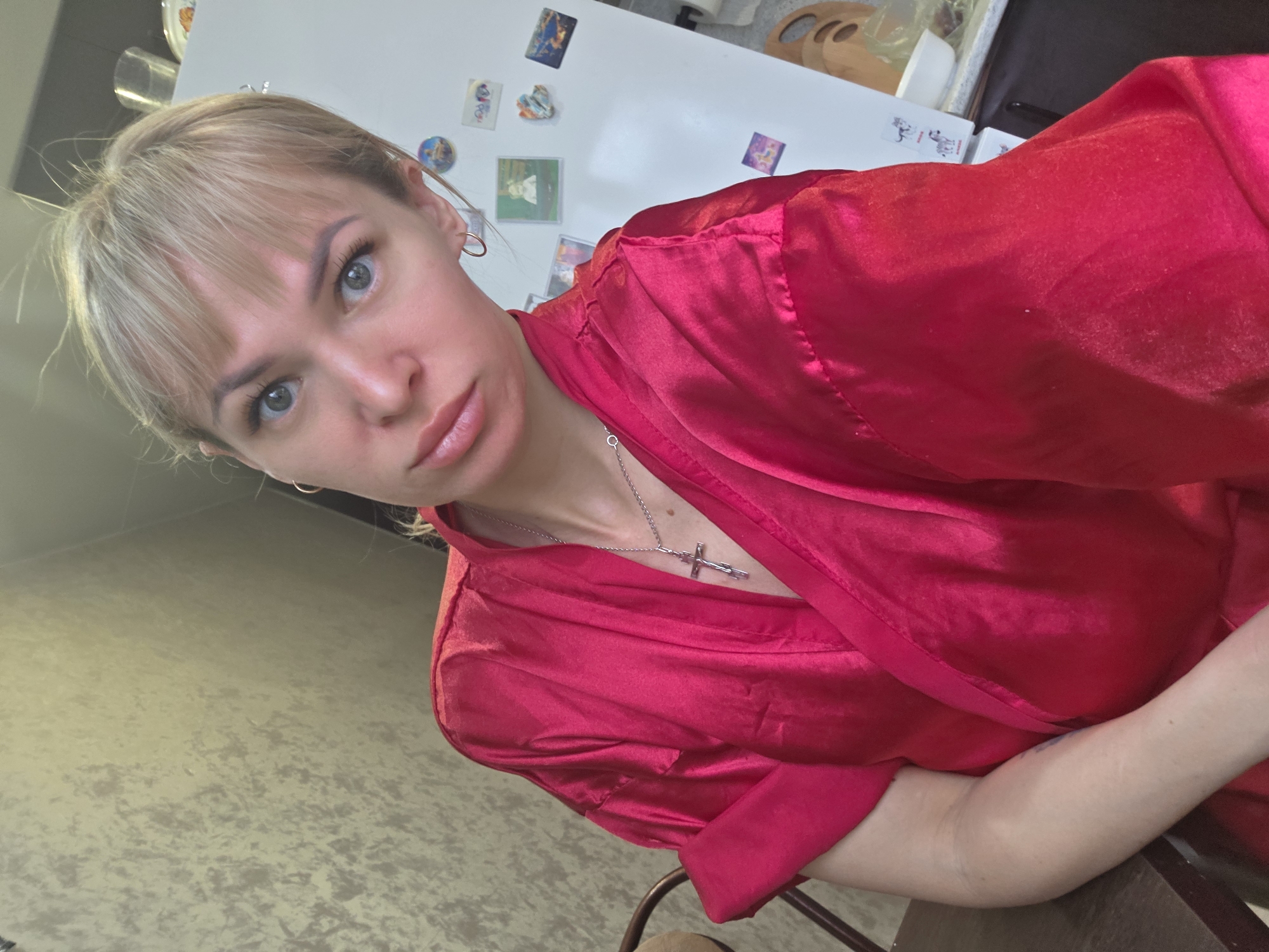 A woman with blonde hair and blue eyes is wearing a red satin robe and a silver cross necklace. She is looking at the camera. The background includes a refrigerator with magnets, a cream-colored wall, and a chair. This image was taken in an indoor location, most likely a residence in the city of Chaikovsky, Russia, based on metadata from other images.