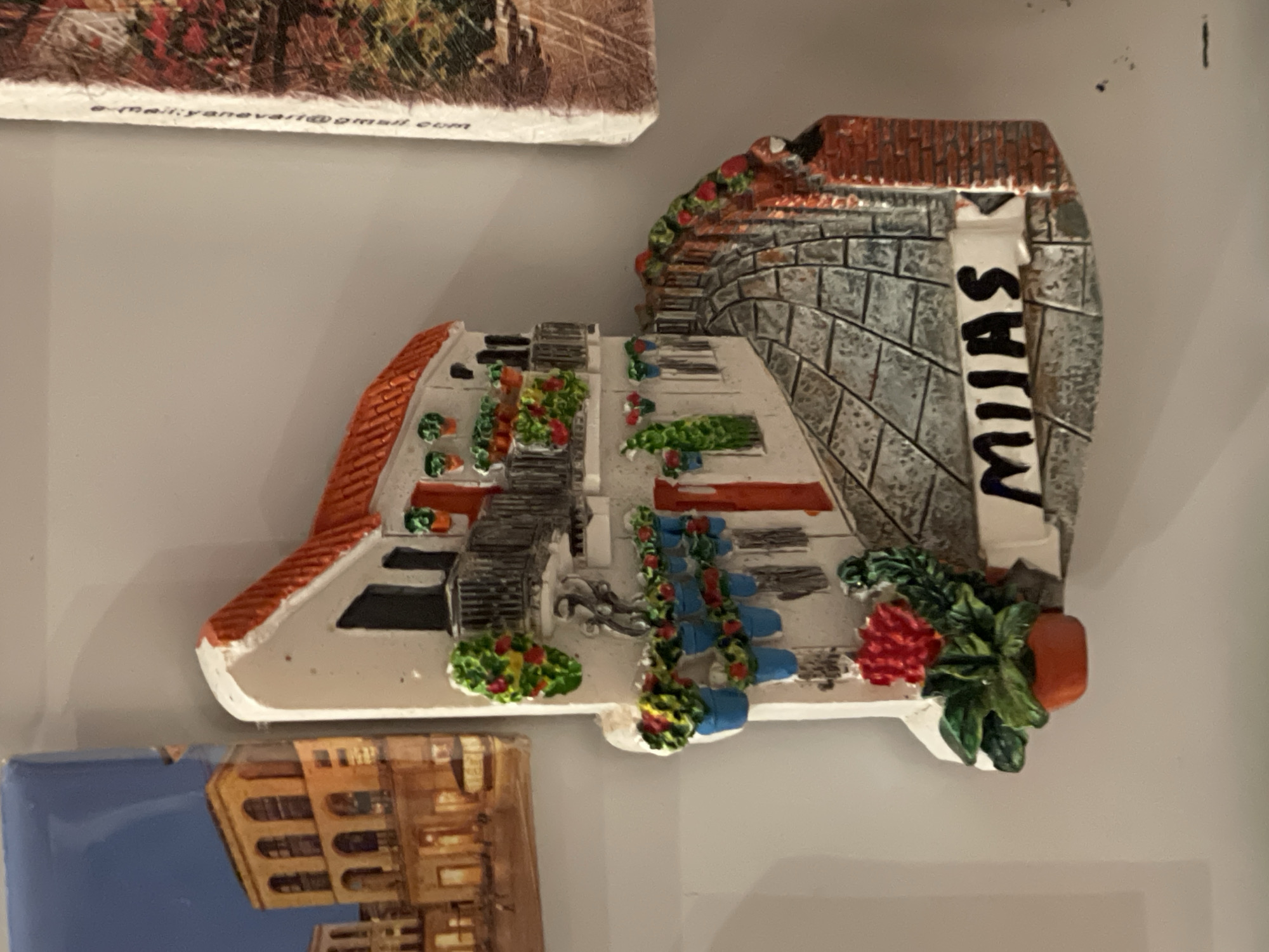 The image shows a decorative magnet of a street scene in Mijas, Spain. The magnet depicts a whitewashed building with a terracotta tiled roof and a stone wall. Flowers are arranged along the stairs and the streets. A vertical banner with the name "MIJAS" is attached to the wall. The magnet is attached to a surface, next to another magnet with a building on it, and another magnet above. There is no evidence to suggest this image is located in Maidstone, United Kingdom.