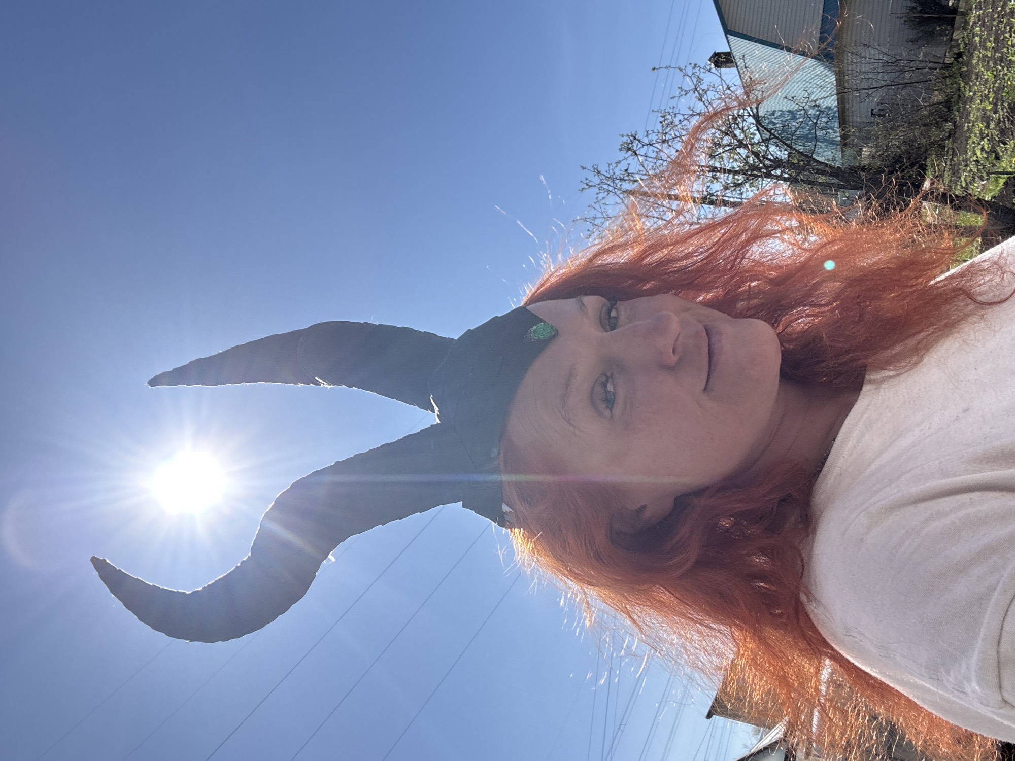 A selfie shows a fair-skinned, red-haired woman wearing a costume headdress with two large black horns that curve upwards. She is positioned in front of a clear blue sky with a bright sun in the upper left, partially obscured by the tip of one horn. Power lines are visible traversing the sky. The background includes glimpses of residential buildings and trees, indicative of an outdoor urban environment. While the city of Chaikovsky, Russia is specified, no features uniquely identify the location, so there's no definitive way to ascertain this based on visual cues alone. The woman is looking down at the camera with a slight smile. She is wearing a light-colored shirt. The angle suggests she's holding the camera above her.