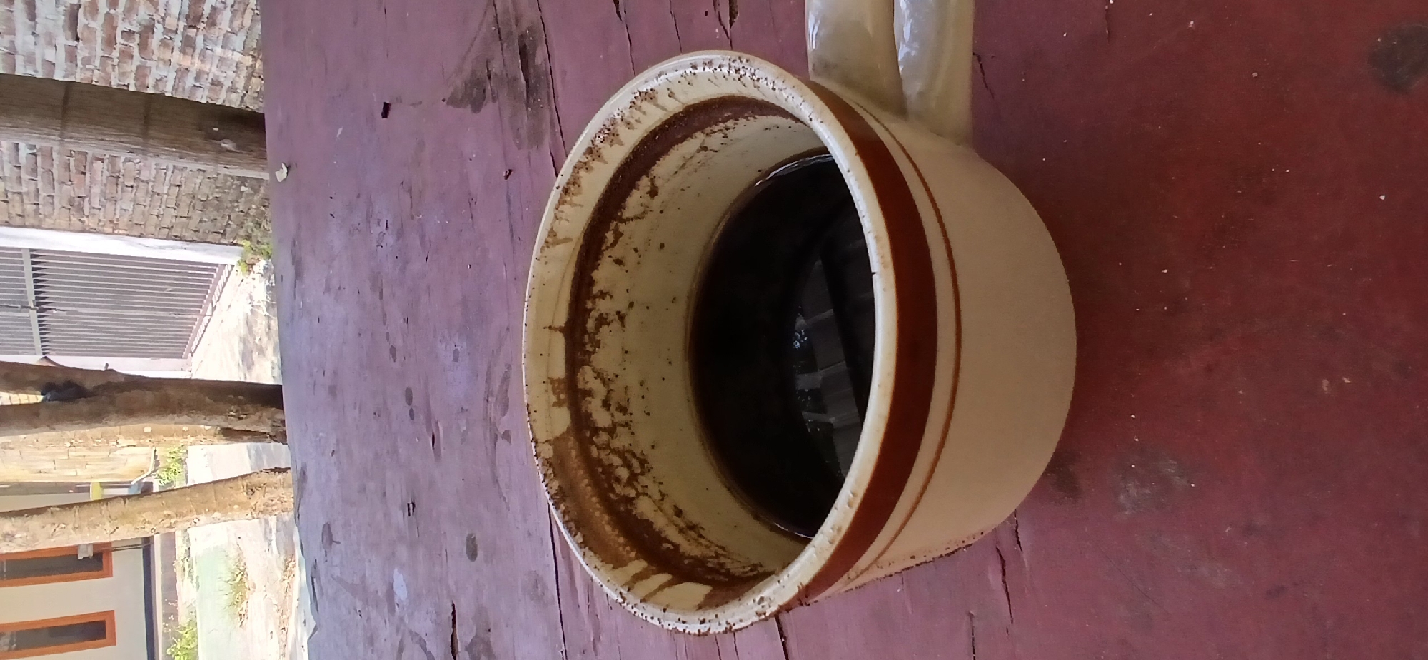 A close-up shot captures a white mug with two brown stripes, filled with dark coffee, resting on a weathered, dark red wooden surface. The rim of the mug is stained with dried coffee residue. In the background, out of focus, are trees, a brick wall, and a metal gate, suggesting an outdoor setting in a residential or urban area. The overall scene evokes a casual, everyday moment, possibly in Pejaten, Indonesia, given the implied context of the location.