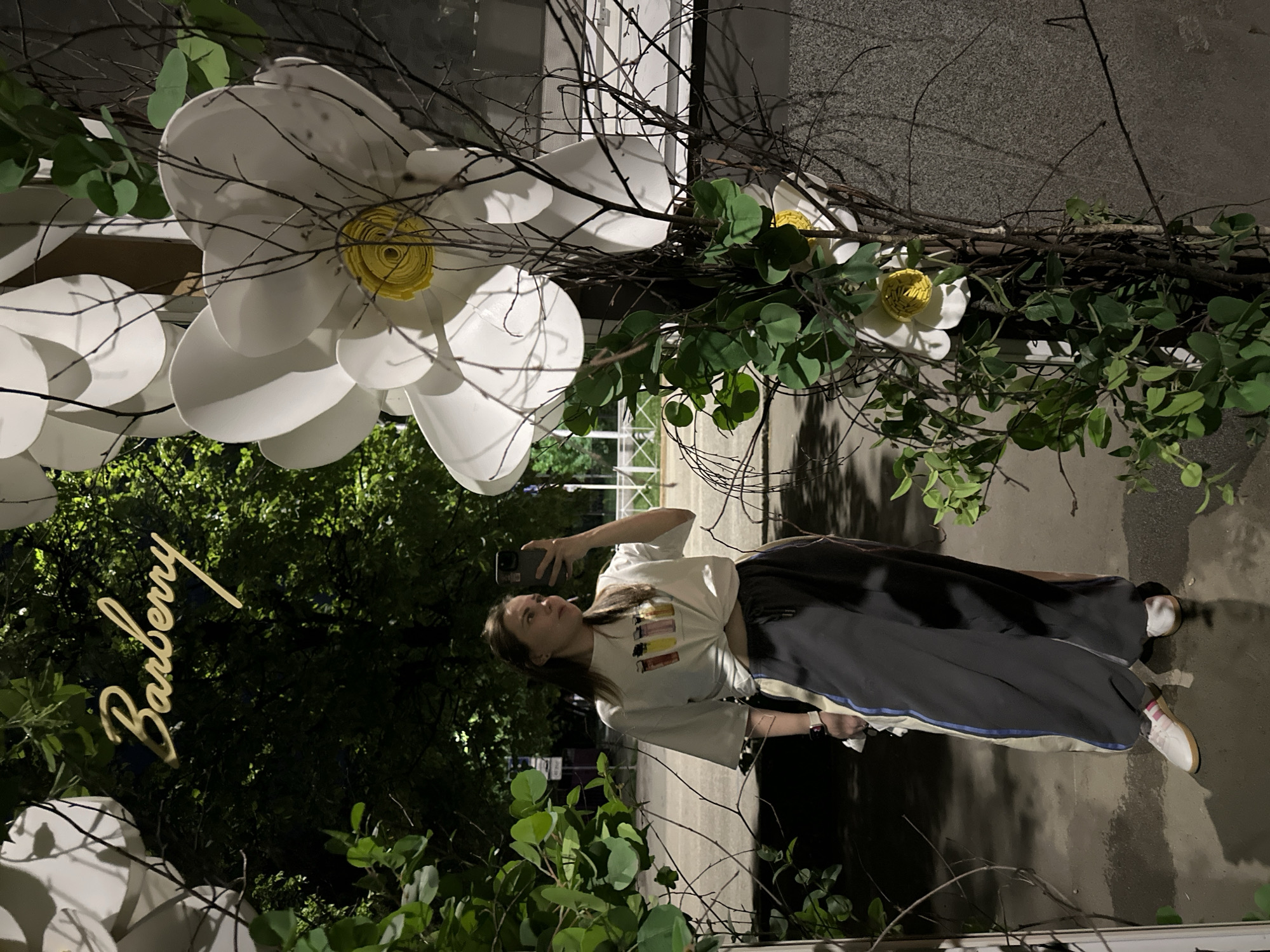 A young woman is photographed in the city of Perm, Russia, in front of an art installation. The installation consists of oversized white flowers and greenery, framing the "Barberry" sign above a park. The woman holds a smartphone, taking a photo or video. She is dressed casually in a white t-shirt and dark sweatpants. The scene is partially illuminated, suggesting it is evening or a low-light environment. The background shows elements of an urban environment, including pavement and foliage.