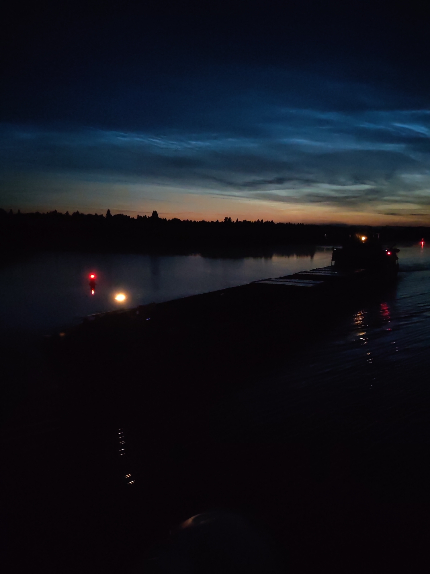 A nighttime view of a barge moving on a body of water, possibly a river or canal, near the city of Kovrizhnovo, Russia. The sky displays a gradient of dark blue at the top transitioning to lighter hues of orange and yellow near the horizon, with wispy clouds illuminated by the fading light. Silhouetted trees line the far bank, creating a dark, indistinct treeline. In the foreground, a barge is partially visible, with its deck appearing dark and wet. A single red navigation light is prominent on a post near the water's edge, with a brighter, yellowish light from the barge itself. The water reflects some of the ambient light and shows ripples. The scene is characterized by low light conditions and a sense of calm.