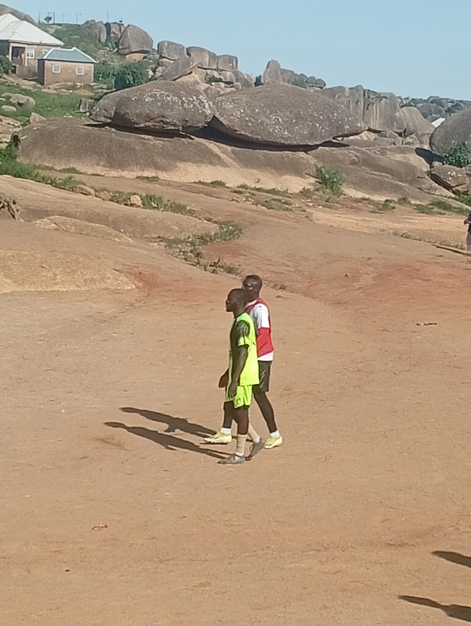 Two young men, likely athletes based on their athletic wear, are walking across a dirt field in a rocky, arid landscape. One man wears a bright green jersey and shorts with black leggings and yellow athletic shoes. The other wears a white and red vest over a black shirt and black shorts with white socks and yellow athletic shoes. They are casting long shadows on the dirt, indicating a bright, sunny day. In the background, large rock formations dominate the scene, with a few small buildings and sparse vegetation visible on the hills. The location appears to be the Shere Hills in Jos, Nigeria, known for its distinctive rock formations and mountainous terrain. The scene captures a casual moment of activity amidst the natural, rugged beauty of the landscape.