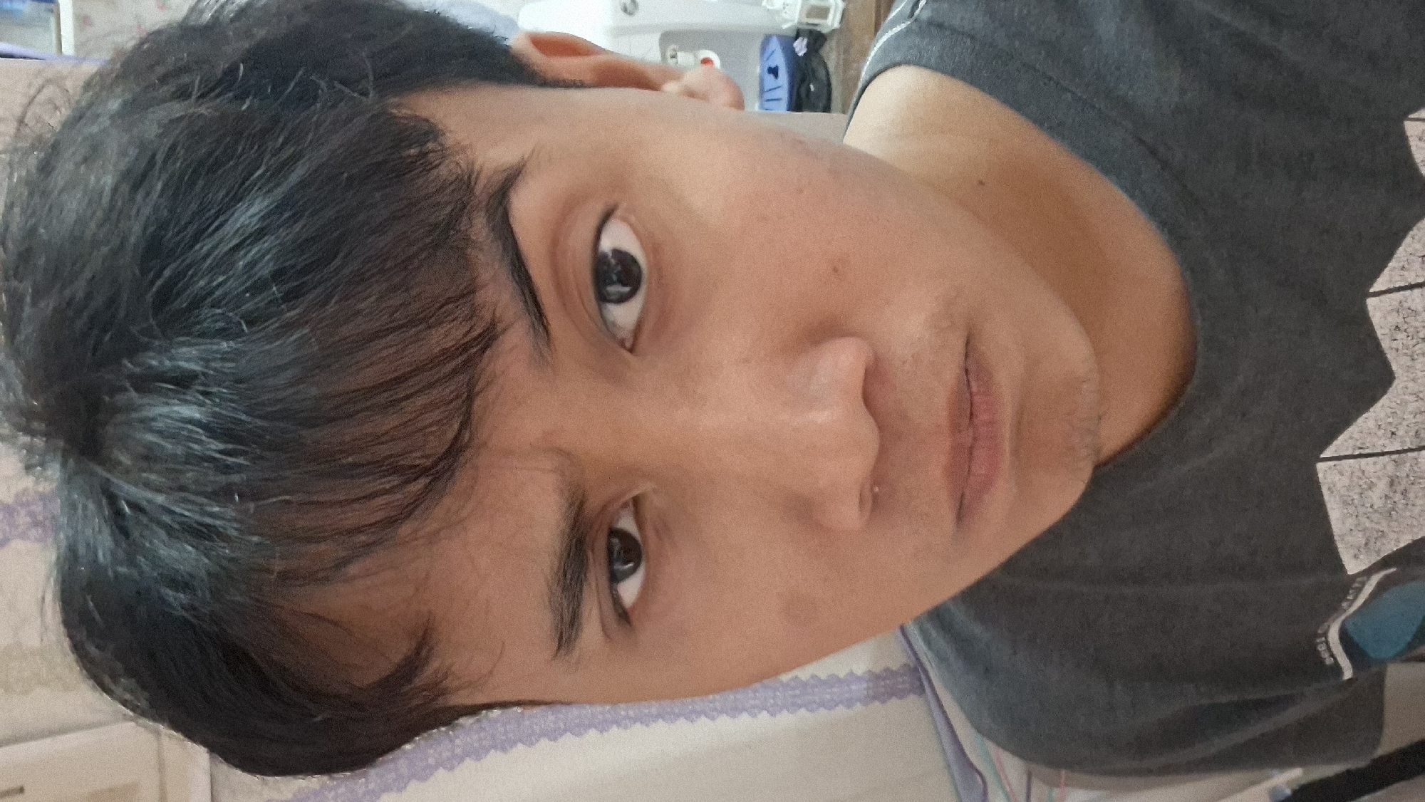 The image, taken in Ciseeng, Indonesia, features a close-up portrait of a young male, appearing to be lying down or reclining, with the photograph oriented sideways (rotated 90 degrees clockwise). He has short, dark, slightly wavy hair that falls across his forehead, and dark brown eyes. Light stubble is visible on his upper lip and chin. His skin tone is medium.
He is wearing a plain dark grey t-shirt, which has a very faint, unidentifiable white graphic or text on the right shoulder area that is too blurry to decipher. His expression is neutral, with his mouth closed and his gaze directed towards the camera.
The background suggests an indoor setting, likely a bedroom or living area. A patterned pillow or bedding with a white and purple design (possibly floral or lace-like) is visible on the left and bottom edges of the frame. In the upper right background, several blurry objects can be seen, including parts of a white structure, possibly furniture or medical equipment, and some blue elements. The lighting is soft and ambient, indicative of indoor conditions, making the specific time of day undeterminable. No clear, readable text is present in the image.