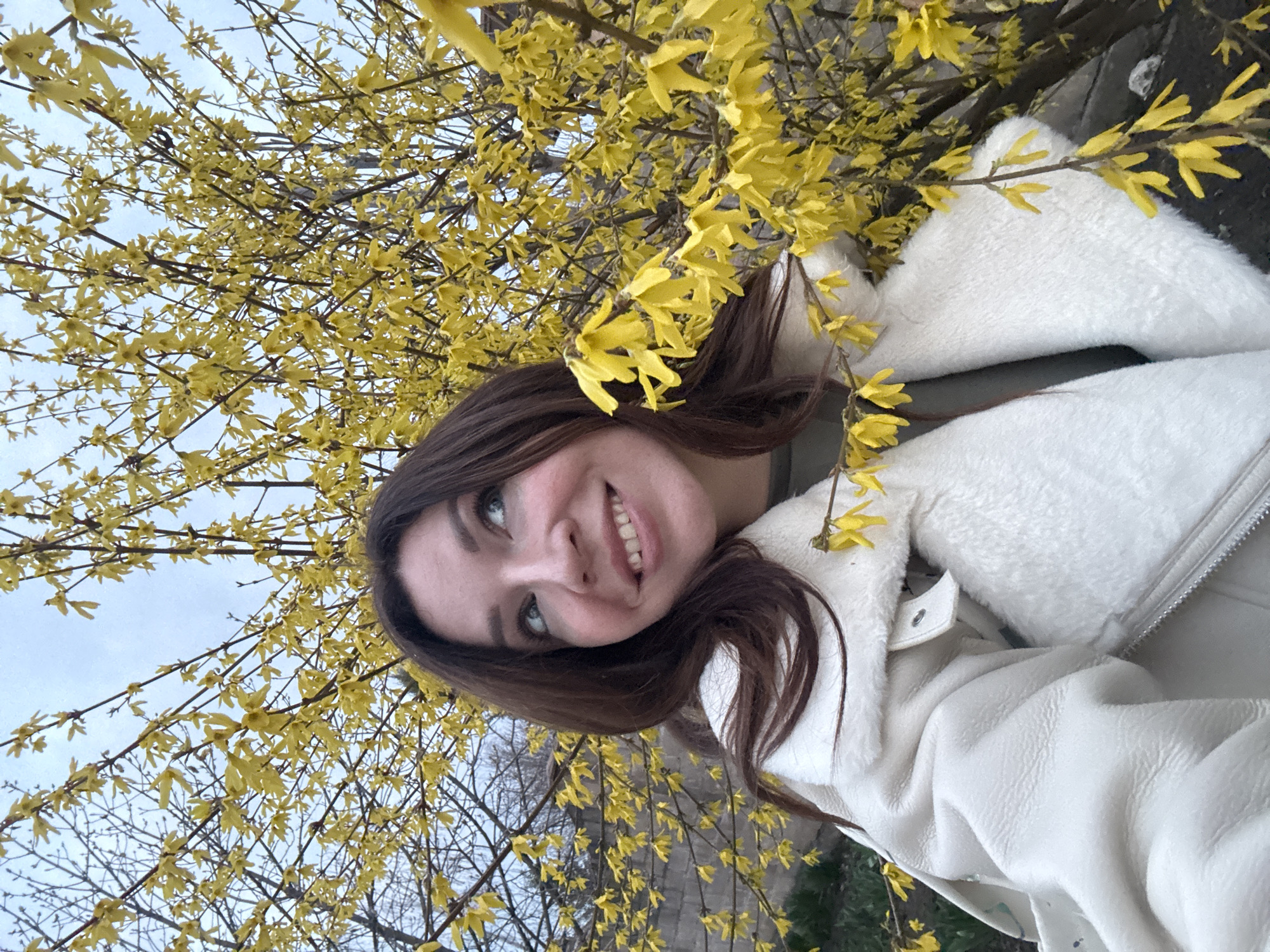 A young woman is smiling and looking upwards in the city of Unknown, Unknown. She is surrounded by a tree with yellow flowers. She is wearing a white jacket. The sky is visible in the background.