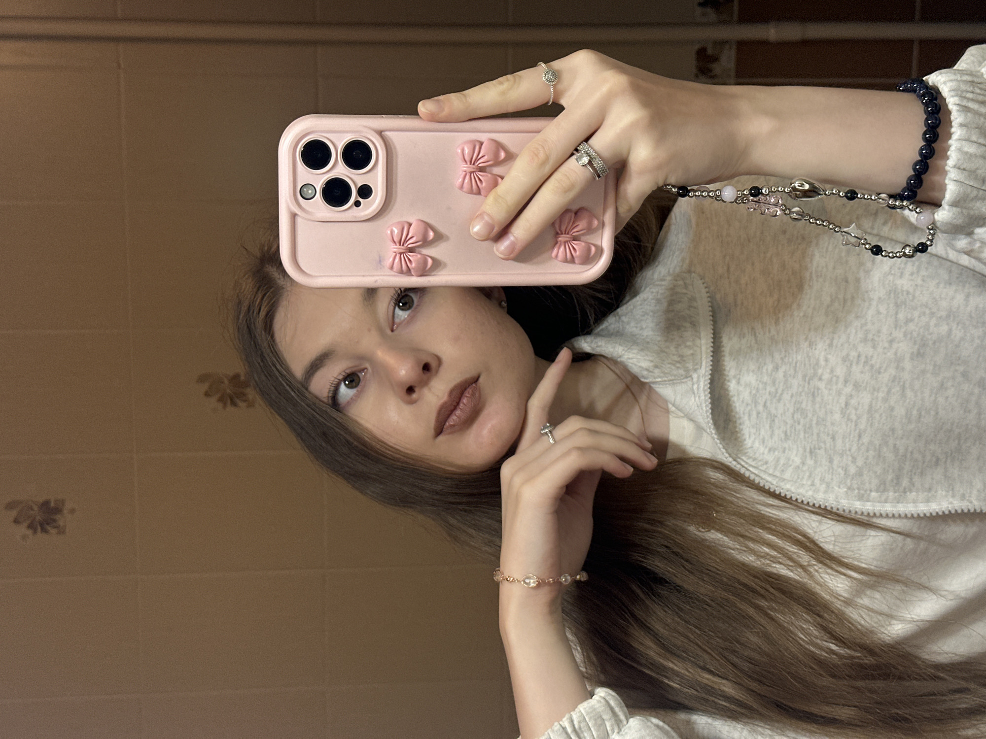 A young woman is taking a selfie in a bathroom. She is holding a pink iPhone with a bow-shaped case in her right hand, and her left hand is positioned near her chin. She is wearing multiple rings on both hands and bracelets on both wrists. She is wearing a light gray zippered sweater. The bathroom has beige-colored floral-patterned tiles. The location is unspecified, but the user has specified it may be in Kazan, Russia.