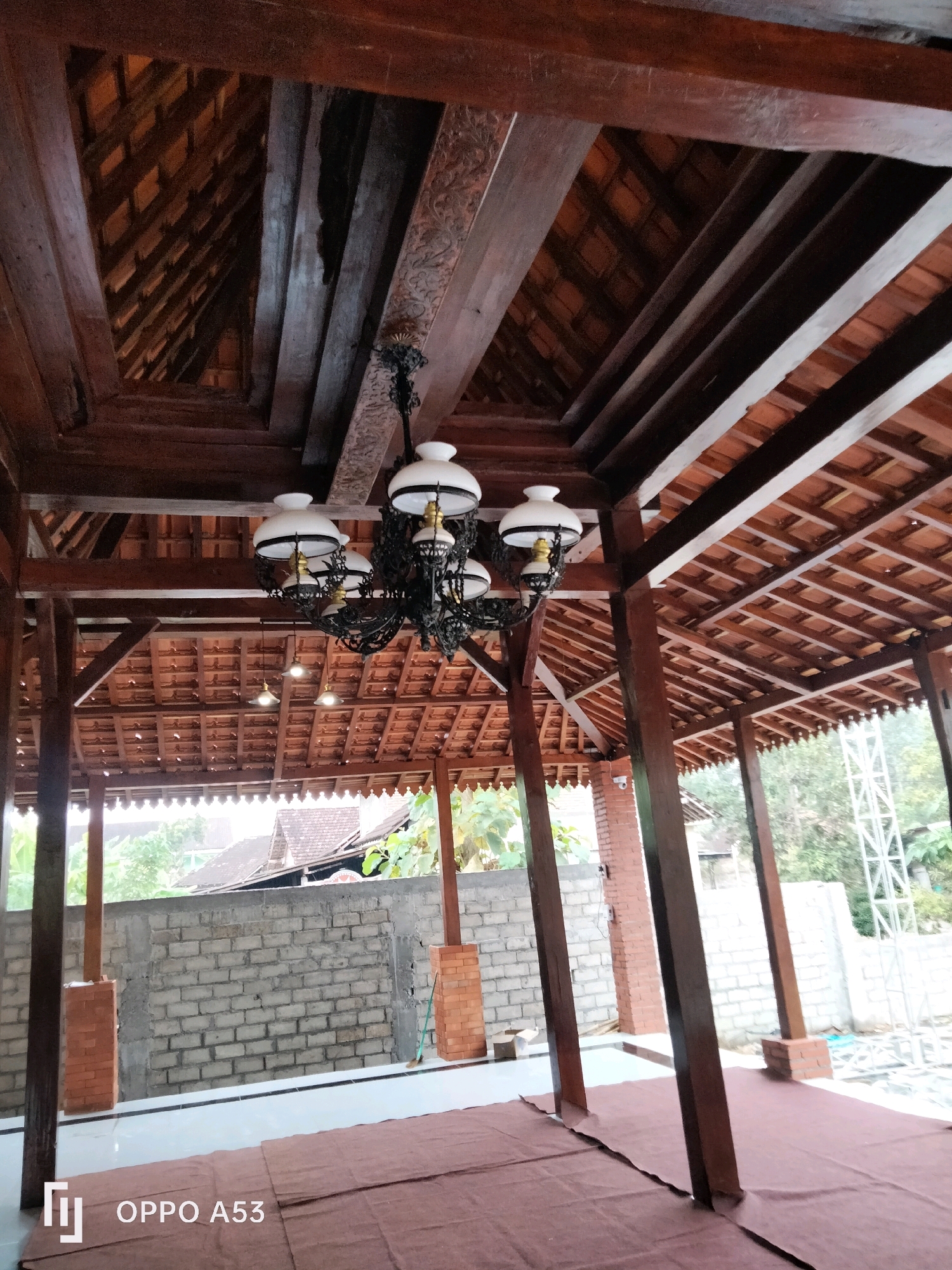 The image shows an open-air structure with a wooden roof and supporting beams. A large, ornate chandelier with multiple white lamps hangs from the ceiling. The roof is constructed with red tiles and intricate wooden detailing. The floor is covered with a brown mat. In the background, a brick wall is visible, and beyond that, trees and a metal structure can be seen. The scene appears to be in a tropical or subtropical outdoor setting. The presence of the "OPPO A53" watermark suggests the image was taken with an OPPO A53 smartphone. Without further context, it is not possible to definitively identify the specific location within Trenggalek, Indonesia.