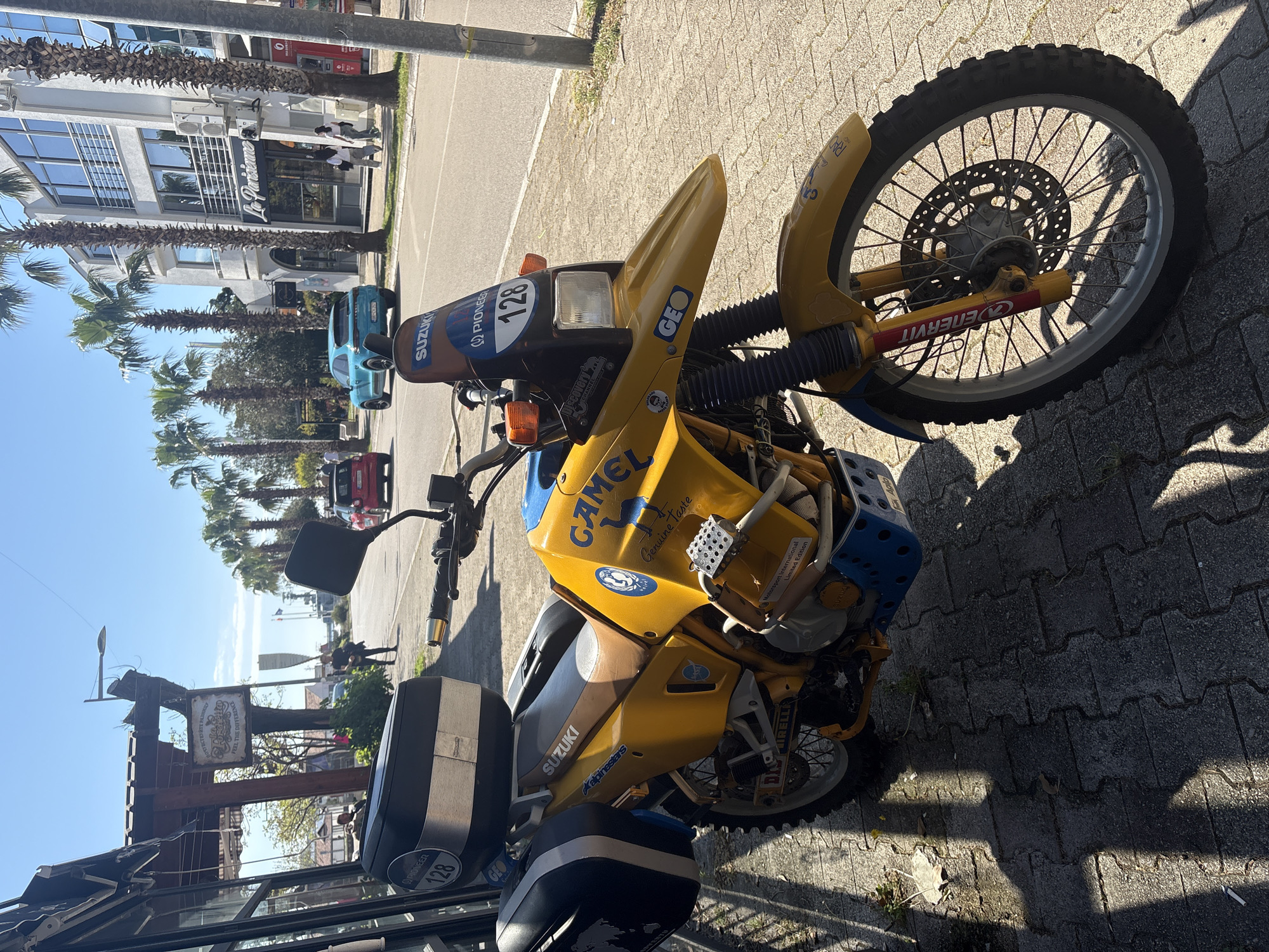 The image shows a yellow and blue Suzuki motorcycle parked on a paved surface. The motorcycle features "CAMEL Genuine Taste" and "Motorsport International Limited Edition" decals. It has panniers and a number plate with "128." There are commercial buildings with palm trees in the background, including one with the name "Le Pariziame." People are walking on the sidewalk in the background. The scene appears to be in Bar, Montenegro.