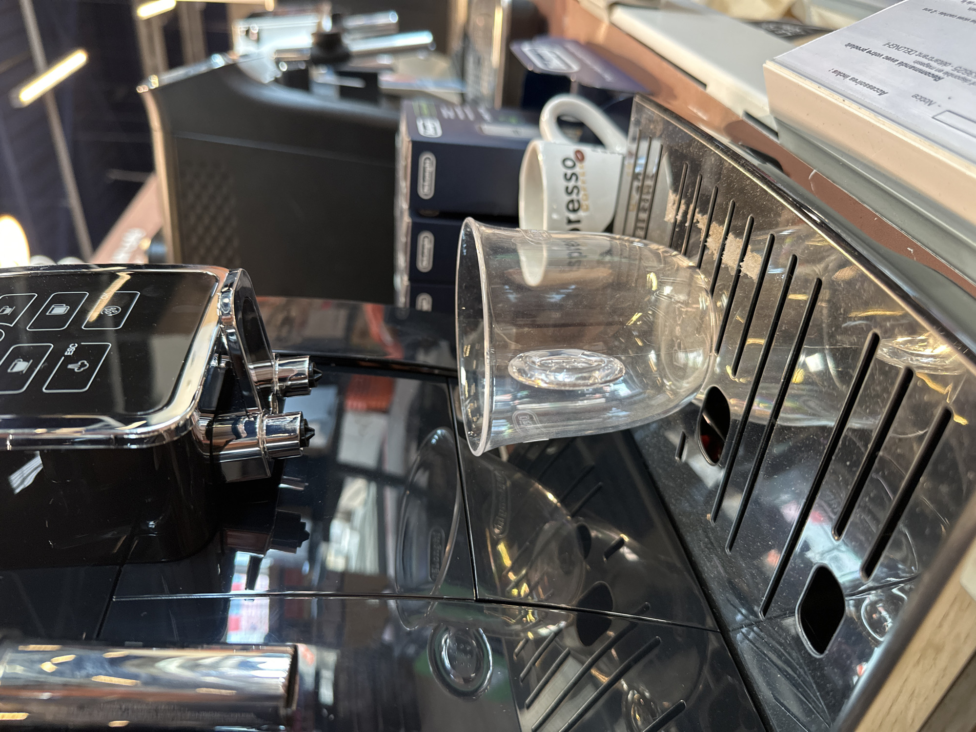 This is a close-up, eye-level shot of a black and chrome espresso machine. The machine has a glossy finish and several buttons with icons. To the right of the machine, there's a clear glass cup, a white coffee mug with the word "spresso" printed on it, and several boxes, one of which has "DELONGHI" printed on it in white text. The background is out of focus but appears to be a domestic or commercial setting with shelving. The lighting suggests it might be indoors during the day. Visible text includes "spresso", "Accessoires inclus:", "Notice", and "DELONGHI".