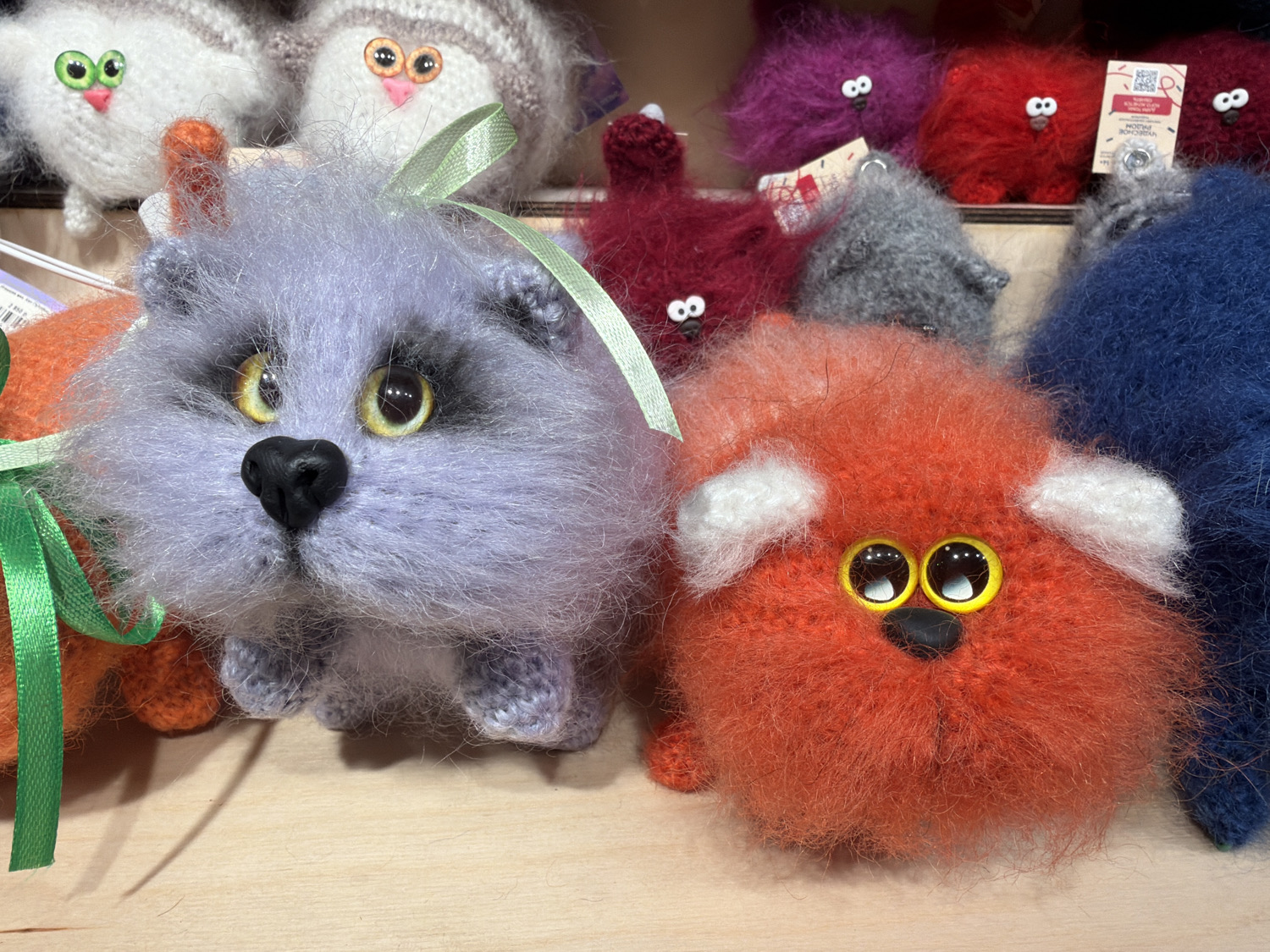 The image presents a close-up view of numerous handmade, fluffy, knitted or crocheted animal-like toys, likely displayed for sale on a light wooden surface. In the immediate foreground, a prominent lavender-colored figure with large, bright yellow-orange eyes and a black heart-shaped nose is visible. It features a light green ribbon tied around its head. To its right, a smaller, vibrant orange toy with large, round yellow and black eyes, a tiny black nose, and distinctive white ear tips is positioned.
Behind these foreground figures, a colorful collection of similar fluffy creatures is arranged. These include white and gray striped toys with simple green or orange knitted eyes, as well as several in rich hues of deep red, magenta, gray, and dark blue, many featuring small, dot-like eyes. The toys all possess a soft, shaggy texture, indicative of fluffy yarn materials.
The scene suggests an indoor setting, likely a craft market stall or a retail shop, given the organized display and the presence of product tags on some of the background items. One such tag, partially visible behind a red toy, displays Cyrillic text that reads "ЧУДО-СОЗДАНИЕ" (Miracle Creation) and "РУЧНАЯ РАБОТА" (Handmade), accompanied by a QR code. The overall impression is one of charming, handcrafted artistry, with a focus on the unique and expressive faces of these whimsical creations. No specific time of day or weather can be determined from the image.