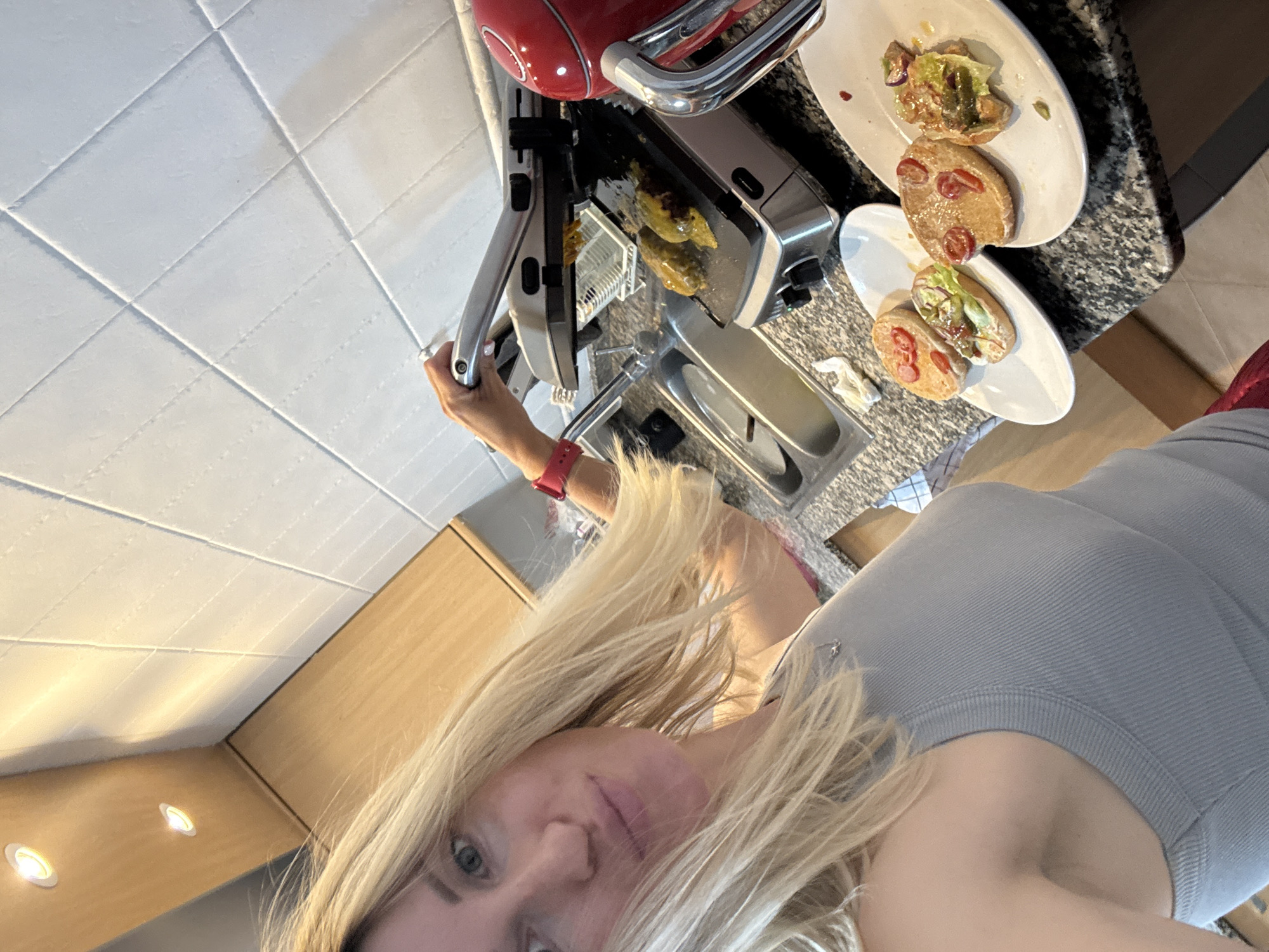 The image shows a woman in a kitchen, likely in a private residence, possibly in València, Spain, given the language requested. The woman, who appears to be Caucasian with long blonde hair, is wearing a light grey tank top and a red watch or bracelet. She seems to be taking a selfie while holding the handle of a red sandwich press, where food is being cooked. Two white plates containing open-faced sandwiches are visible on a grey countertop with a marble-like pattern. The kitchen also features white tiled walls and wooden cabinets with integrated lighting.