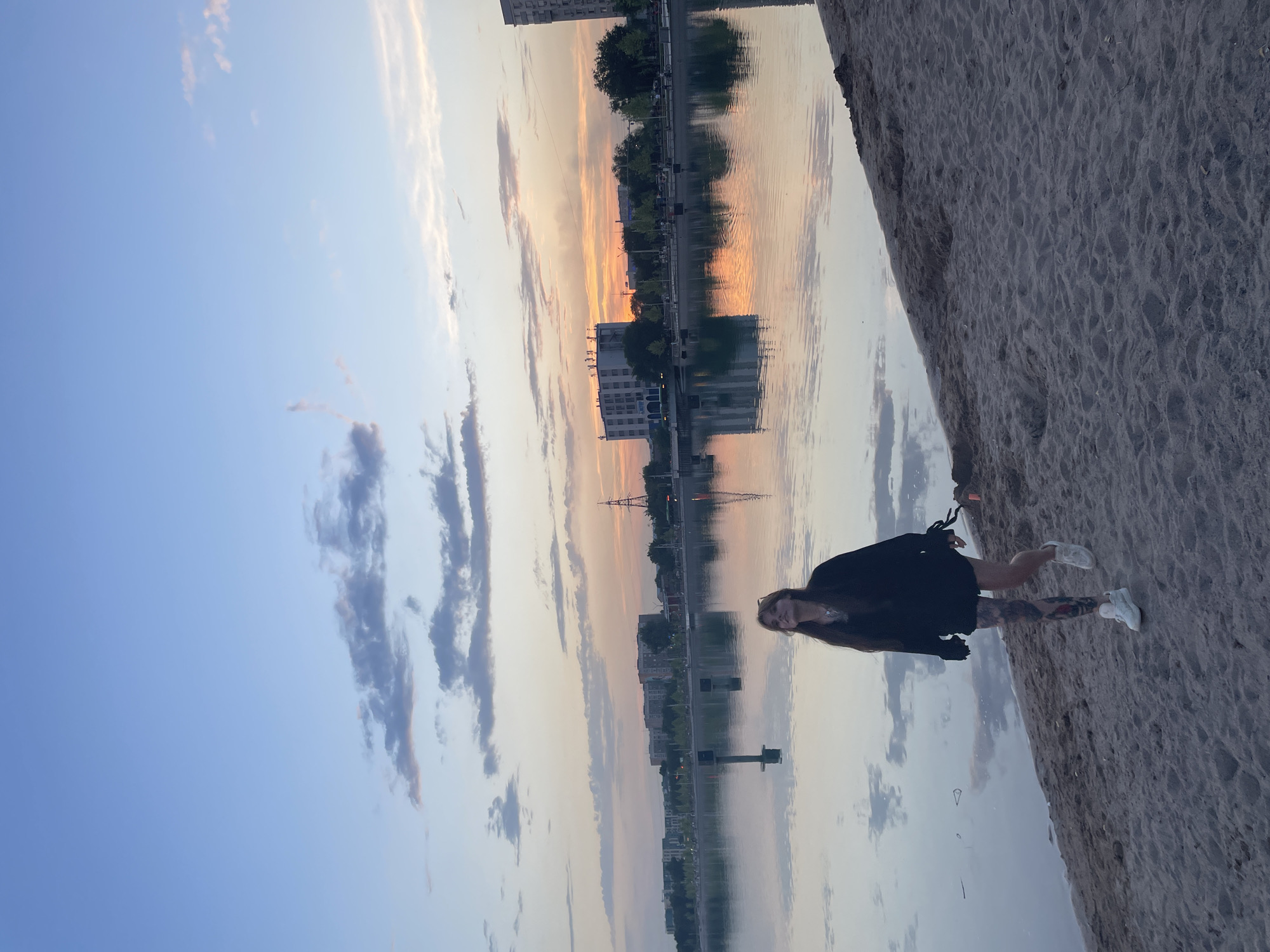 A young woman with tattoos on her legs walks along a sandy shore next to a calm body of water. She is wearing a black, oversized hoodie and white sneakers. The sky is a gradient of blue and orange, with scattered clouds, indicating either sunrise or sunset. Across the water, there is a city skyline with buildings and trees. The water reflects the sky and the buildings. The scene appears to be located in Almaty, Kazakhstan, possibly along the Kok-Tobe hill embankment or near the city's fountains on an embankment, given the presence of a cityscape, water, and recreational areas.