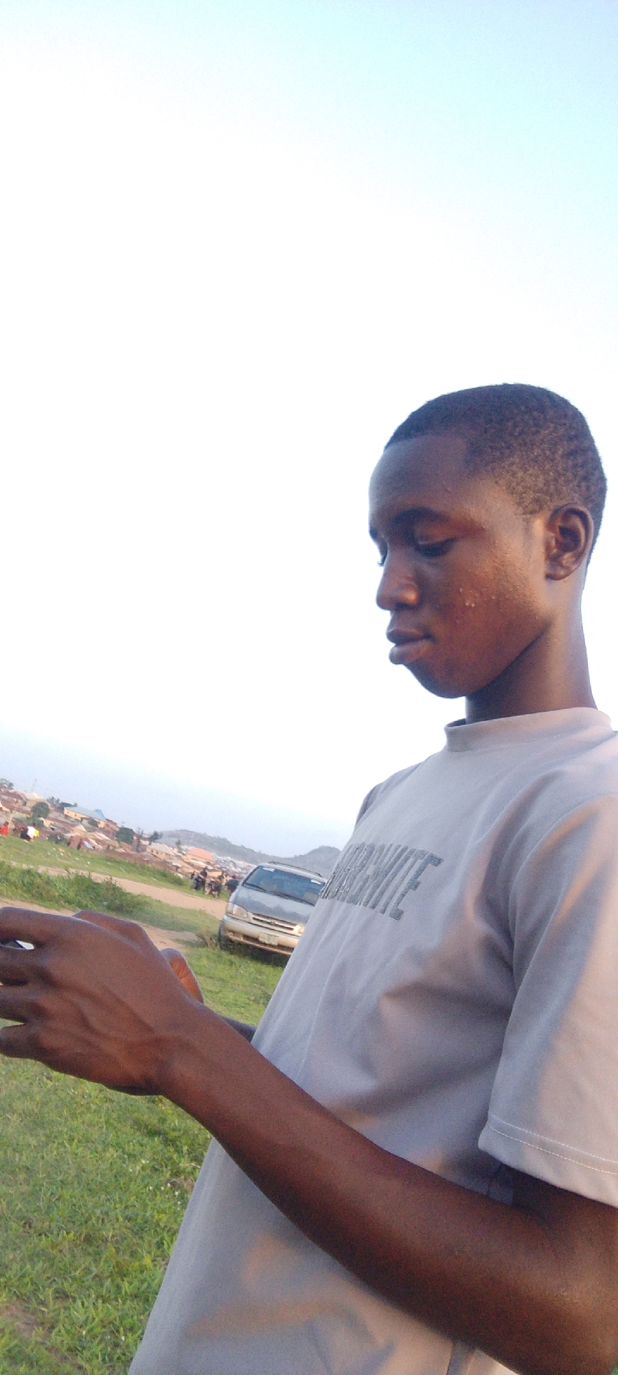 A medium shot captures a young Black man outdoors. The subject, positioned on the right, wears a light-colored T-shirt with a visible graphic text across the chest. He gazes down at an object in his hands. The lower section of the frame shows a patch of green grass, hinting at a natural environment. In the background, a road is visible with a silver car parked beside it. Several people are present nearby. A cluster of buildings can be seen in the distance, indicating an urban environment. The sky is a light blue. The location appears to be on the outskirts of a populated area, as evidenced by the nearby buildings and activity. The man's dark skin and short hair are noticeable features.