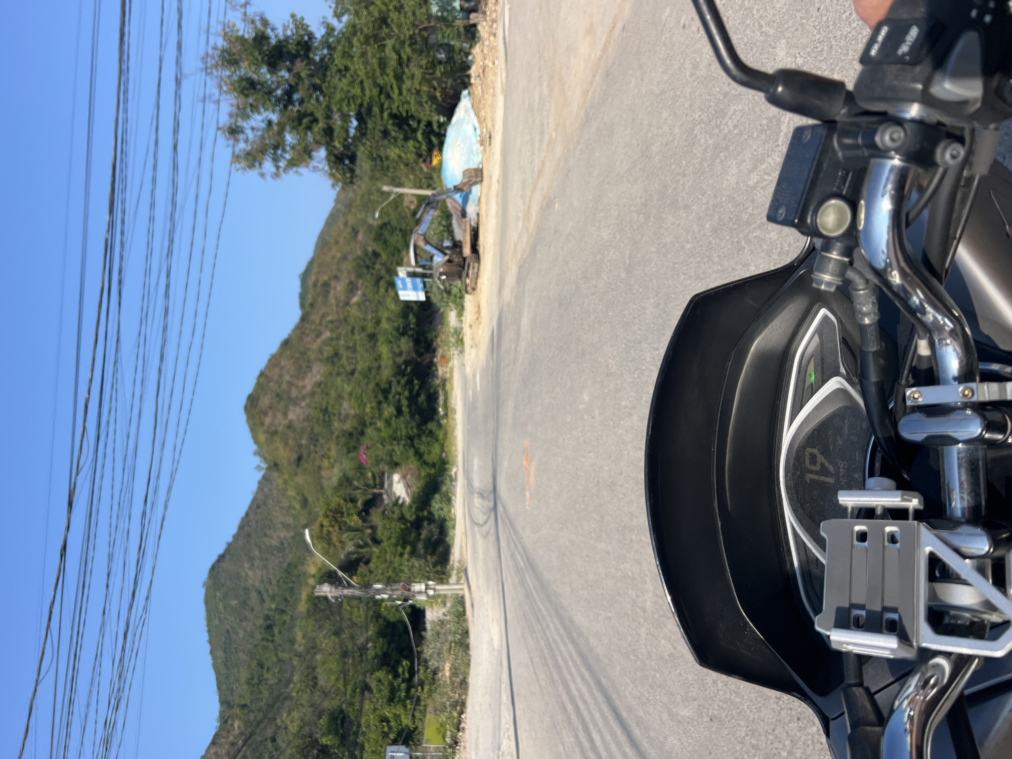 This vertical image captures a first-person perspective from a motorcycle or scooter, indicating a journey through a scenic, mountainous region, likely Bắc Nha Trang, Vietnam. The bottom right portion of the frame is dominated by the vehicle's black dashboard, handlebars, and a chrome phone mount. The digital display on the dashboard is visible, showing the number "19" and partially visible "12:48," which likely represent the speed and current time.
Spanning the left and upper parts of the frame, an asphalt road stretches into the distance under a clear, bright sky. The road is bordered by an unpaved shoulder where an excavator or small digger is parked, suggesting ongoing construction or earthworks. Behind the machinery, a mound of earth is covered with a light blue tarp. A blue and white sign, too blurry to discern text, stands beside the excavator.
The background features lush, green, tree-covered hills or mountains, rising prominently against a vibrant, cloudless blue sky. Numerous dark power lines extend horizontally across the upper expanse of the sky, adding a man-made element to the natural landscape. The strong sunlight casts distinct shadows, confirming it is daytime, likely mid-morning to afternoon. The scene conveys a bright and clear day, showcasing both natural beauty and evidence of human development along a roadside route.