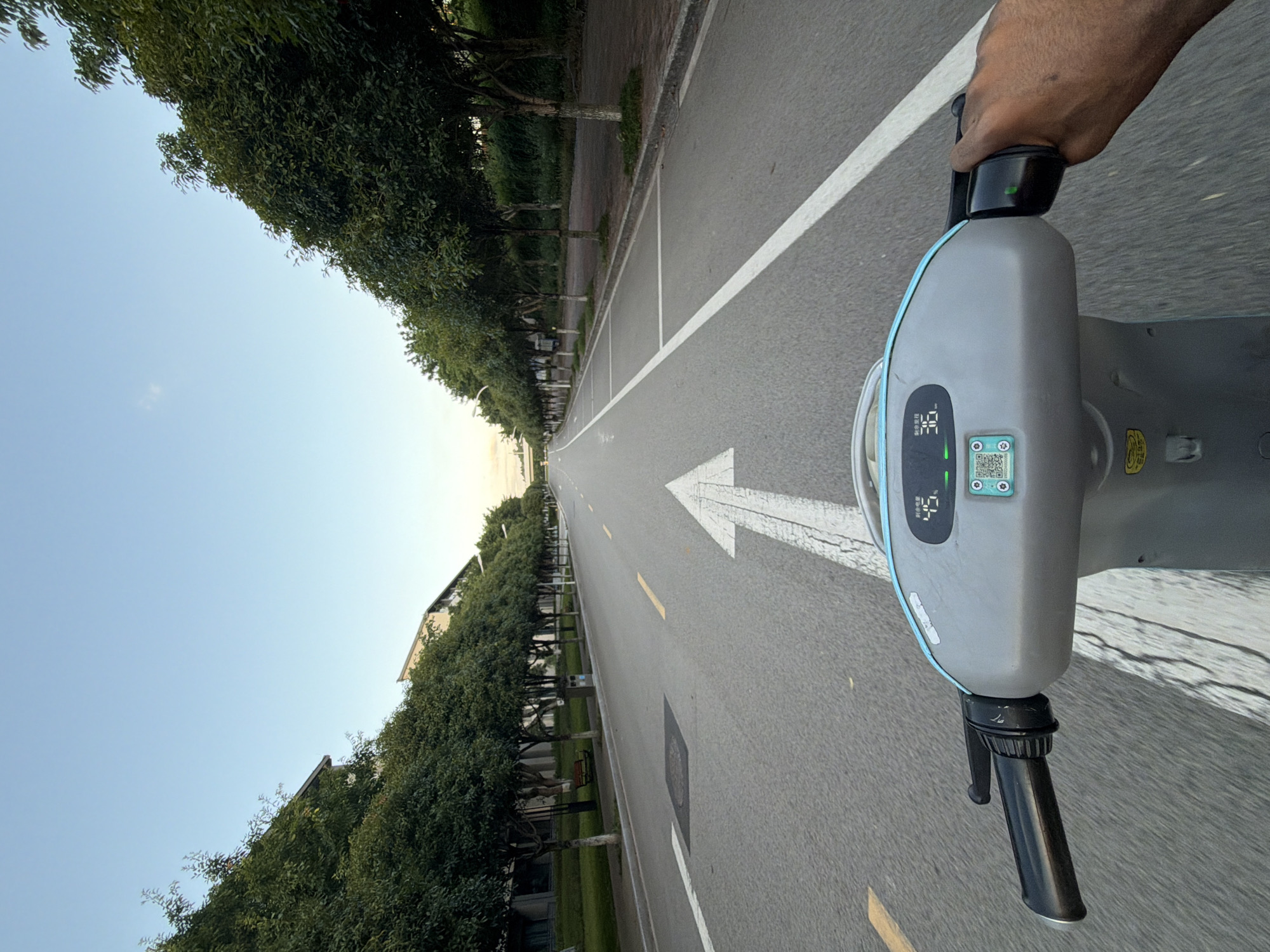 This image depicts a first-person perspective of someone riding an electric scooter on a road lined with trees. The road is asphalt with a white arrow painted in the center, indicating the direction of travel. On the scooter's dashboard, a digital display shows "45" and "36", likely representing speed and battery level or distance. On the right side of the dashboard, a QR code is visible, along with some Chinese characters. A hand is visible gripping the scooter's handlebar on the right. The environment appears to be a campus or park area with buildings visible in the background, partially obscured by trees. The sky is clear and blue, suggesting it is daytime. The scene is likely in the city of Jiangjin, China, given the context of similar images from that region.