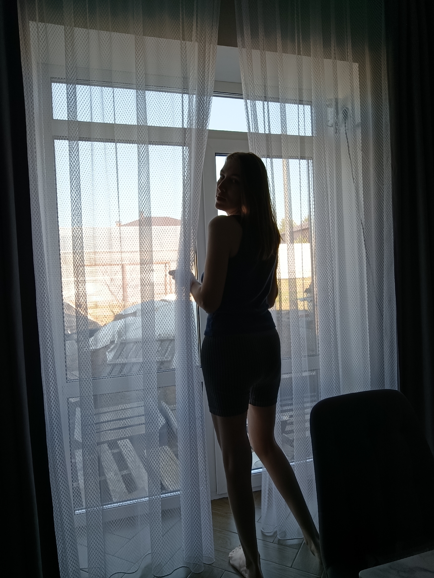 The image shows a woman standing by a window in a room. She is wearing a dark tank top and shorts. The window has a white lace curtain and a view of what appears to be a residential area with houses and a fence outside. A dark curtain is visible on the right side of the window. In the foreground, a dark chair is partially visible. The setting appears to be indoors, possibly in an apartment. It is difficult to confirm the location as Chaikovsky, Russia without further details or context.