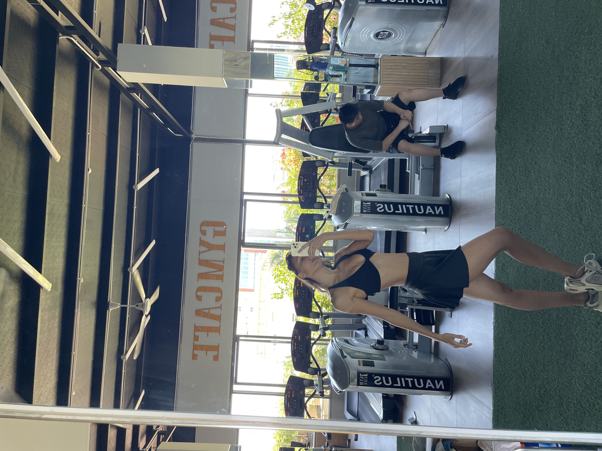 A woman in athletic wear poses for a mirror selfie in a gym. She is wearing a black sports bra and a black skirt. In the background, several treadmills are visible, along with other gym equipment. Another person, a man, is seated on a piece of equipment, looking down. The gym has large windows offering a view of trees and buildings, suggesting an urban or suburban setting. The sign above the treadmills reads "GYMCAFE" in large, orange letters. The flooring is a combination of wood and green turf. The overall lighting suggests it is daytime. The location is identified as Ham Thang Ward, Vietnam.