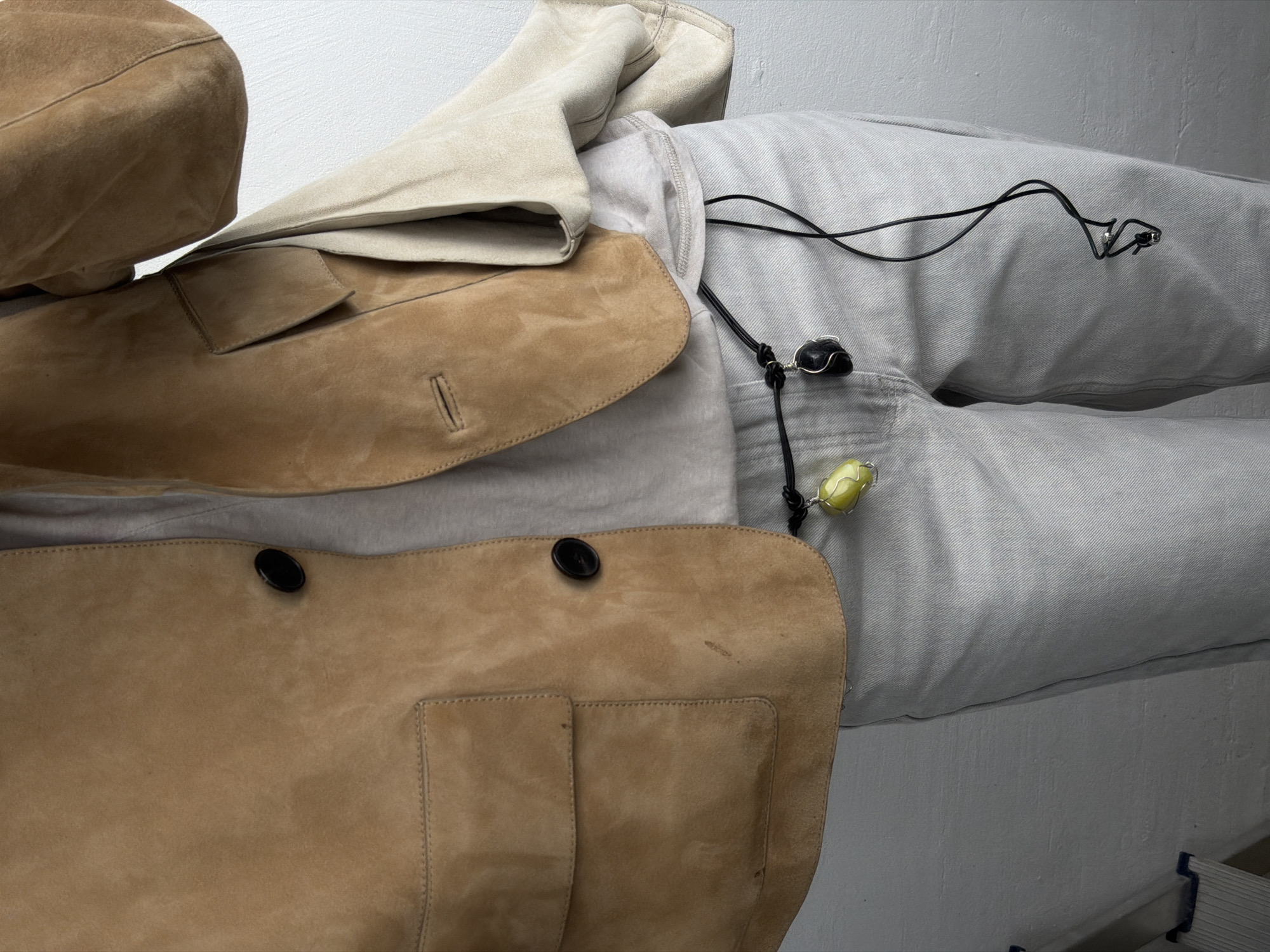 The image shows an overhead view of a selection of clothing items. A light brown suede jacket with visible pockets and black buttons is prominent. A beige beret rests near the jacket. The ensemble is placed on top of light grey sweatpants with a black necklace featuring a light green stone. The scene is indoors with a light grey background, possibly a wall or a table. There's no discernible information linking this to Khimki, Russia.