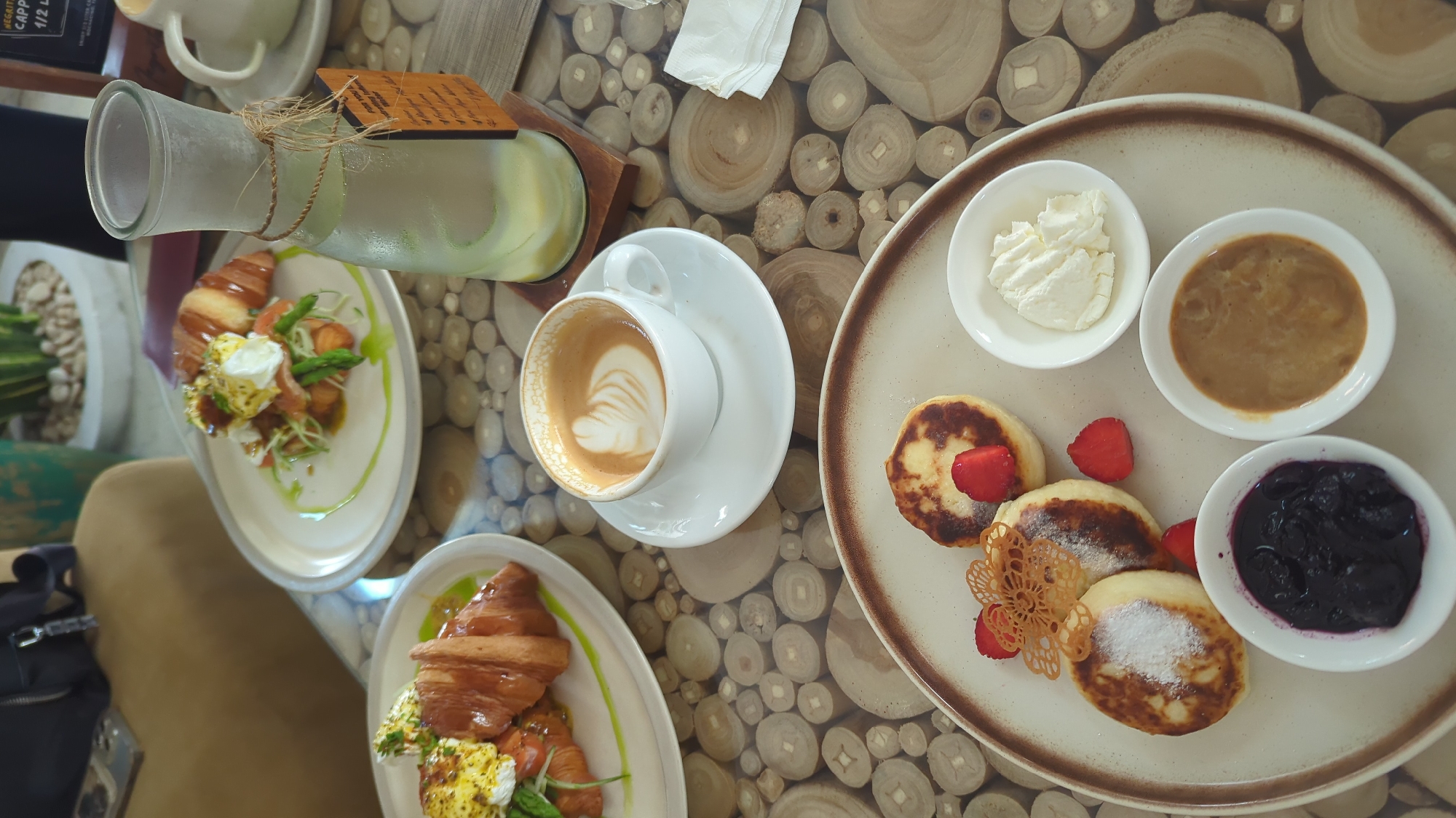 An overhead shot captures a vibrant breakfast or brunch spread laid out on a distinctive table crafted from numerous small, circular wooden cross-sections. The scene depicts a relaxed dining experience, likely at a cafe in North Kuta, Indonesia, with natural daylight suggesting a morning or midday meal.
In the foreground, a large round plate holds several small, golden-brown pancakes, some dusted with powdered sugar, and garnished with fresh strawberry halves and an intricate, delicate wafer decoration. Alongside, three small white bowls contain accompaniments: one with white cream (possibly whipped cream or fresh cheese), another with a light brown, creamy sauce (like caramel or peanut butter), and a third with dark purple blueberry jam.
To the left, a white ceramic cup of coffee with elegant latte art rests on a saucer, positioned near the center of the table. Behind the coffee cup, a menu card is partially visible with the text "VEGITABL..." and "CAPPUCCINO". Further left, two identical plates are visible, each featuring a large, flaky croissant accompanied by a serving of what appears to be scrambled or poached eggs, green asparagus spears, and fresh greens, all drizzled with a light green sauce.
A clear glass bottle, with a frosted appearance and tied with natural twine and a small wooden tag, holds a light green beverage, possibly a refreshing infused water or lemonade. Text on the wooden tag is partially obscured and unreadable. In the background, hints of potted plants with white pebbles suggest an outdoor or open-air setting. A person's knee and part of a dark bag are visible in the lower left, indicating a diner seated at the table.