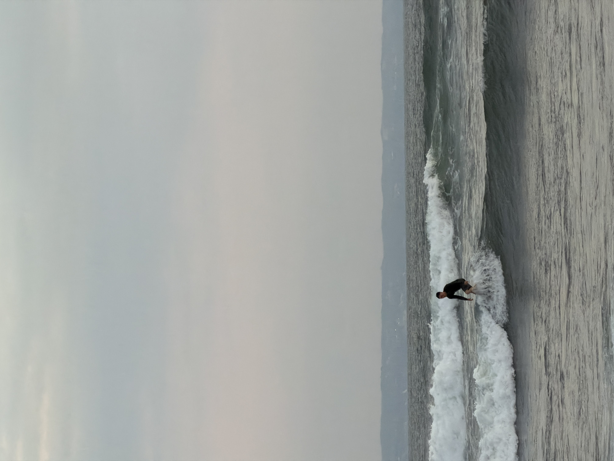 A surfer in a black wetsuit rides a wave in the ocean. The surfer is positioned on the right side of the frame, facing left. The sky is overcast and a distant shoreline with hazy hills is visible on the horizon. The ocean is dark gray with white crests on the waves. The scene depicts a surfing activity in the waters off North Kuta, Indonesia.