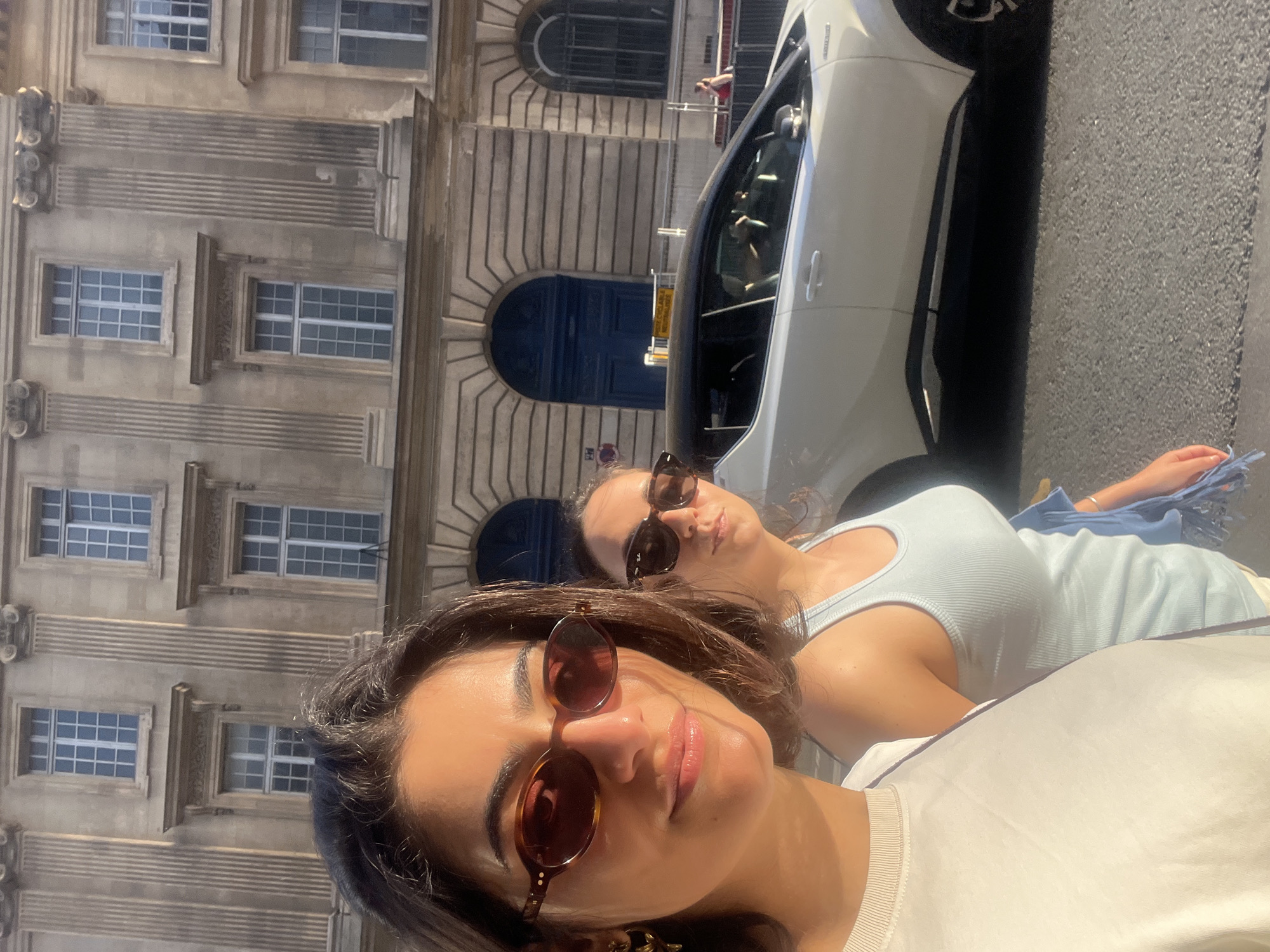 Two women, wearing sunglasses, pose in front of a car and a building in Paris, France. The building has multiple stories, with several rectangular windows on each floor. Arched doorways are visible. The car is white. The street surface is dark.