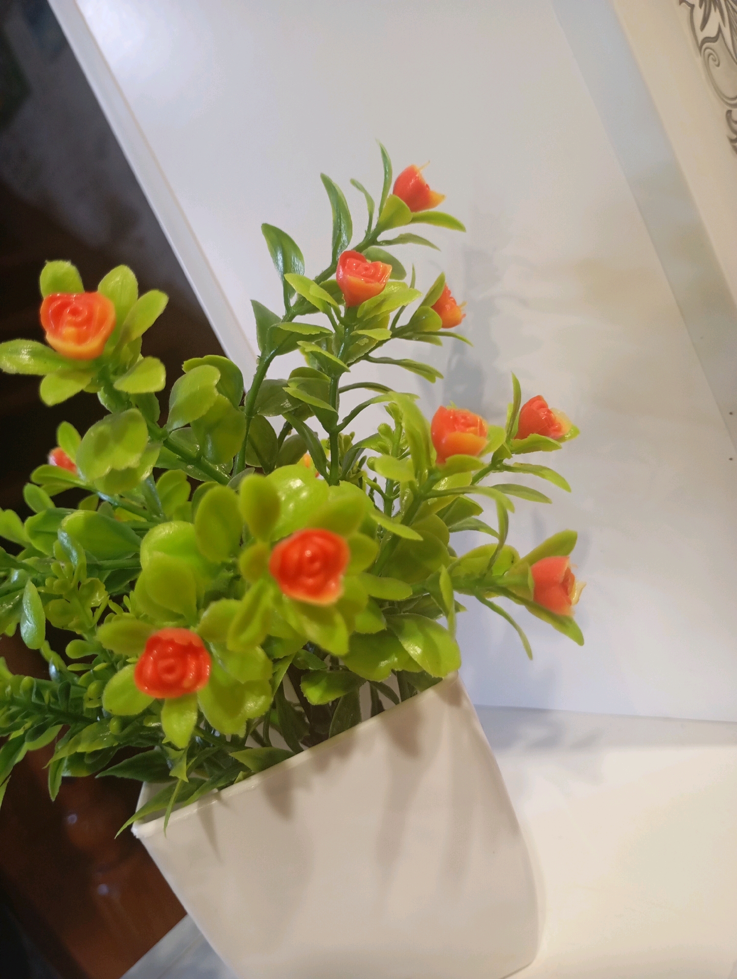 A potted artificial plant with green leaves and small orange flowers sits in a white vase. The vase is positioned on a flat surface, likely a shelf or table. The background is divided diagonally, with a dark, indistinct area on the left and a lighter, plain surface on the right. The lighting is artificial and casts soft shadows. There is no visible indication of the city of Фоки, Russia.
