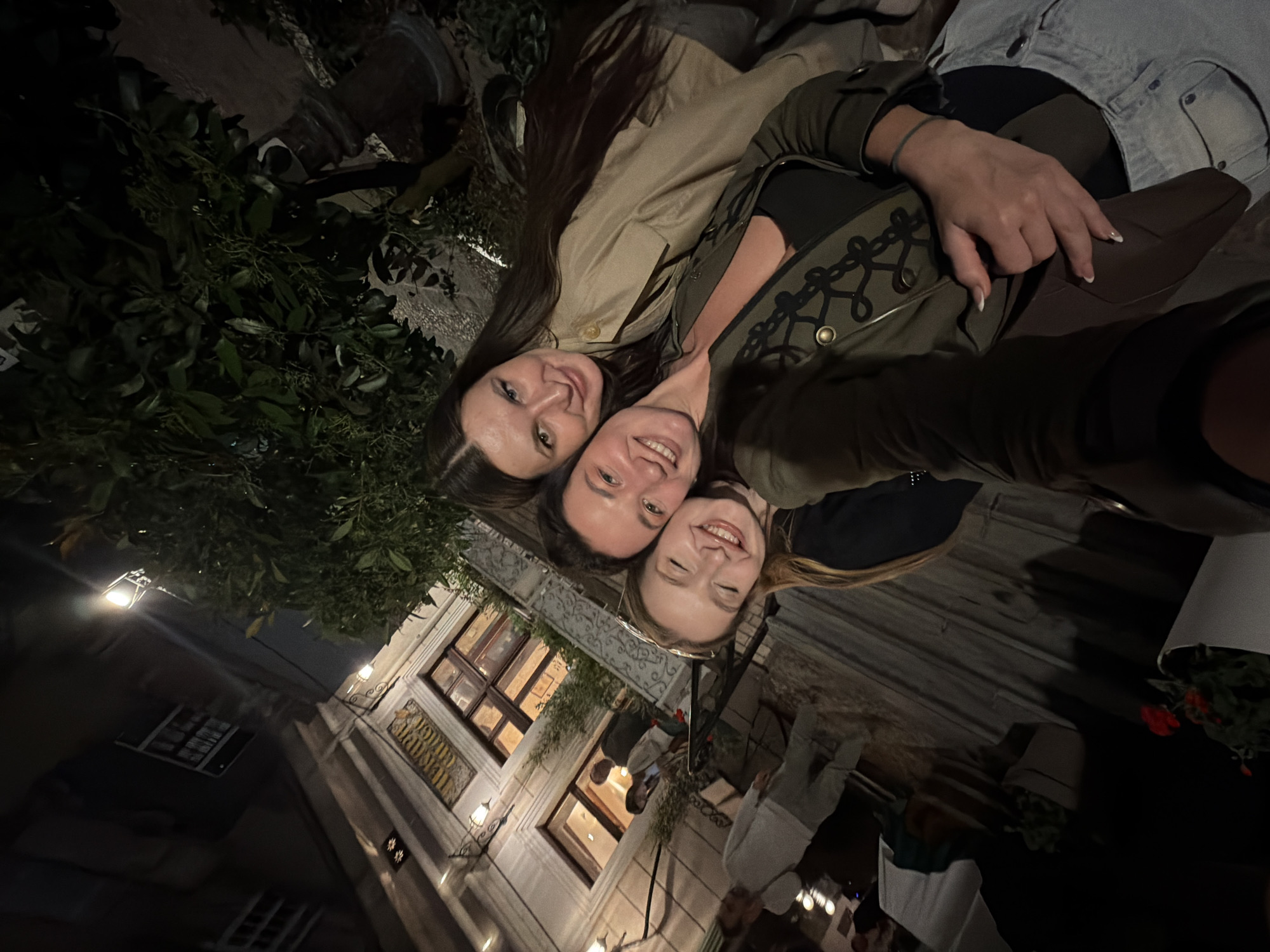 Three women are taking a selfie outdoors at night, with architectural features suggesting a European setting. The woman on the left has long dark hair and is wearing a light tan jacket. The woman in the middle has dark hair and is wearing an olive green jacket with decorative embellishments. The woman on the right has blonde hair. A building with lit windows and ornate decorations is visible in the background. Lush greenery is present to the left of the women. Tables and figures that appear to be dining patrons are also visible in the background. The location is Belgrade, Serbia.