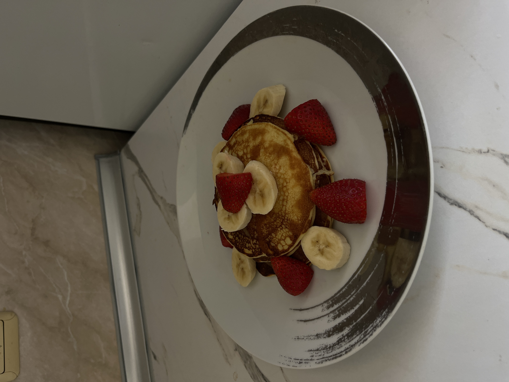 The image depicts a plate of pancakes adorned with sliced bananas and strawberries. The plate has a white surface with a decorative gray and black rim. The pancakes are stacked in the center of the plate. The plate is placed on a light-colored marble countertop, and a portion of a white cabinet is visible in the background. The location cannot be determined from the image alone.