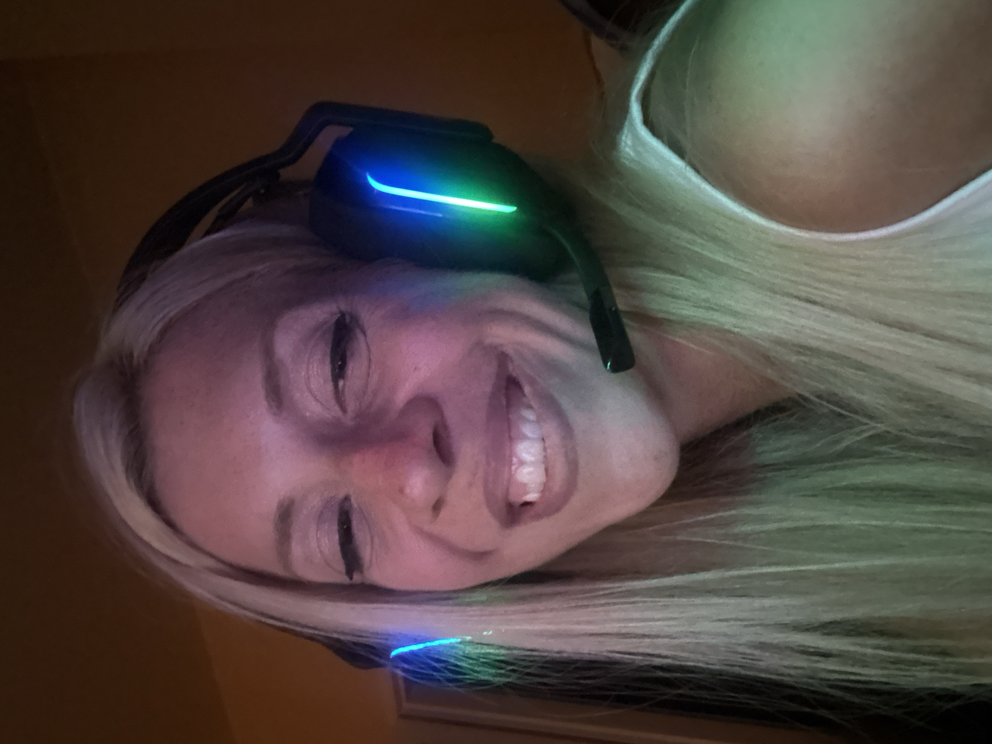 The image shows a close-up of a woman wearing black headphones with blue and green LED lights. She has blonde hair and is smiling. The background is dark and out of focus, with some warm lighting. The image does not contain any elements that would allow for the identification of a location in Pazardzhik, Bulgaria.