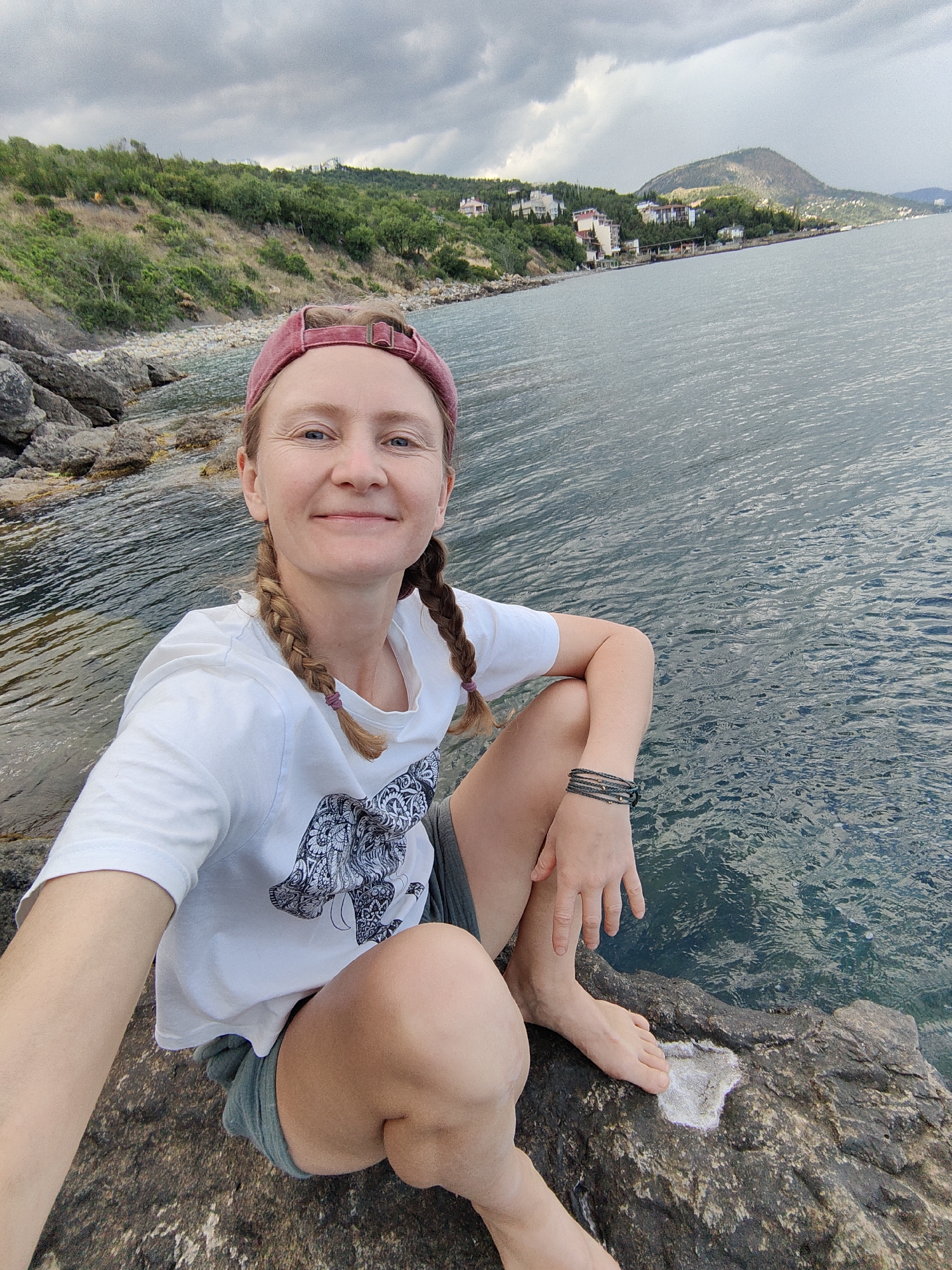 A woman with light brown hair in two braids, wearing a red baseball cap, a white t-shirt with a black elephant graphic, and grey shorts, sits on a rocky shoreline. She is smiling at the camera, with her left arm bent and her hand resting on her knee. She has a dark bracelet on her left wrist. The ocean is to her right, with gentle waves. Behind her, a hilly landscape with trees and buildings is visible along the coast. The sky is cloudy. The scene appears to be in Utios, Ukraine.