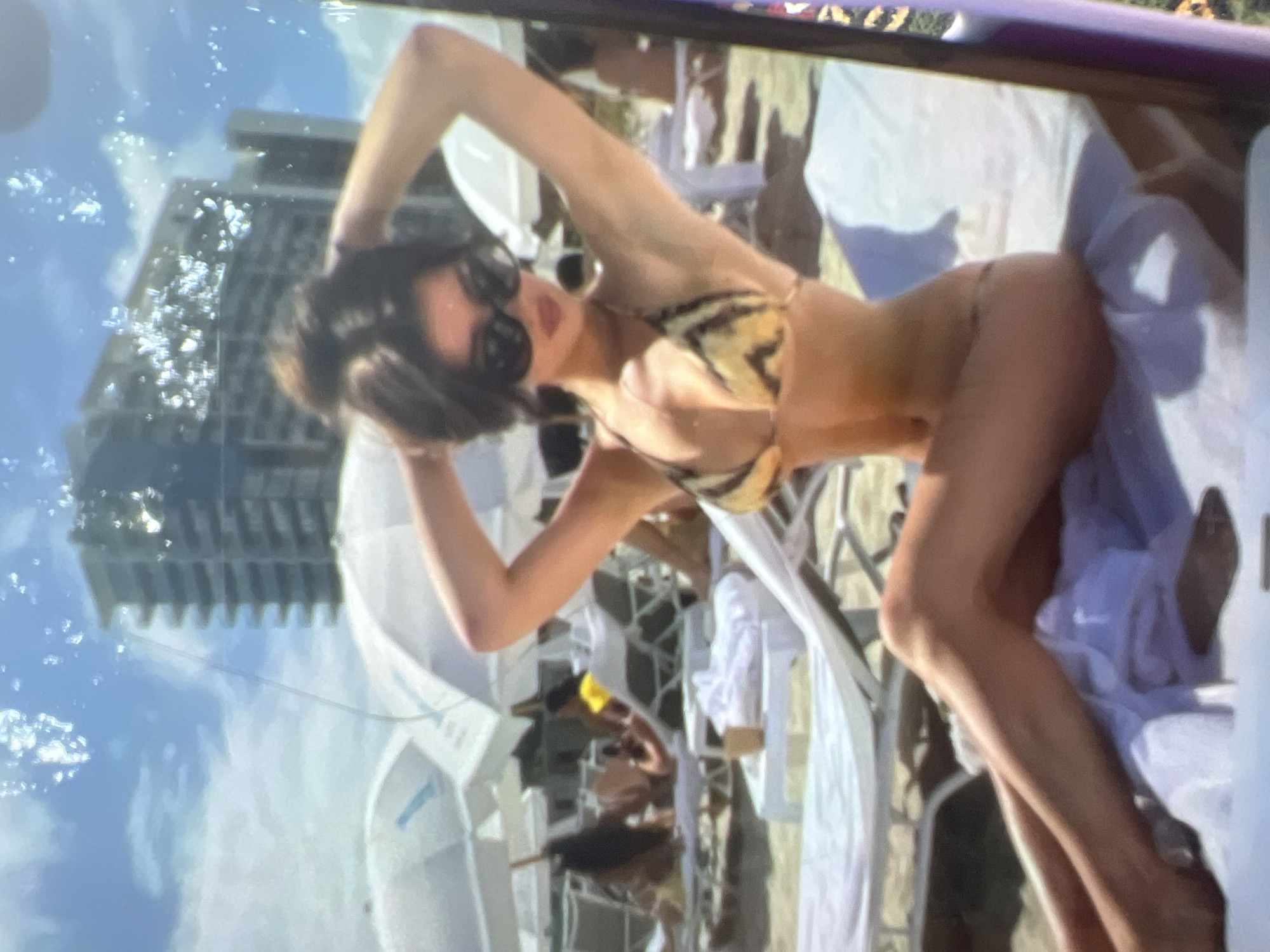 The image shows a woman in a bikini sunbathing on a beach. She is lying on a lounge chair, with her hands behind her head. She is wearing sunglasses and a striped bikini top. In the background, there are beach umbrellas, lounge chairs, and a building. The scene appears to be a sunny day at the beach. The image quality is poor and there are visible artifacts, possibly from scanning or a damaged screen. The location of Kara-Dıykan, Kyrgyzstan is not identifiable from the visual information in the image.