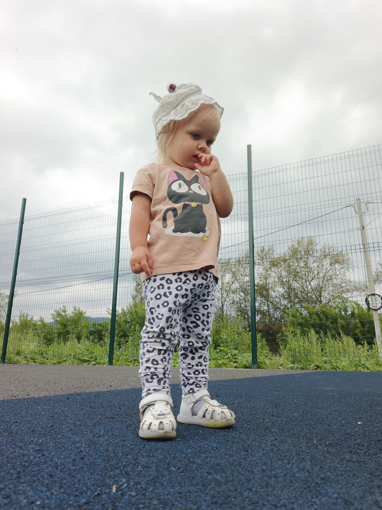 A low-angle full shot shows a young Caucasian child, wearing a white sun hat with a purple pom-pom, a peach-colored t-shirt with a black cat graphic, and white leopard-print pants, standing on a blue rubberized surface in an outdoor recreational area. The child is looking down with their right index finger in their mouth. Behind the child, a green metal fence with wire mesh separates the recreational area from a grassy embankment with trees and a cloudy sky. The overall scene suggests a park or playground setting.