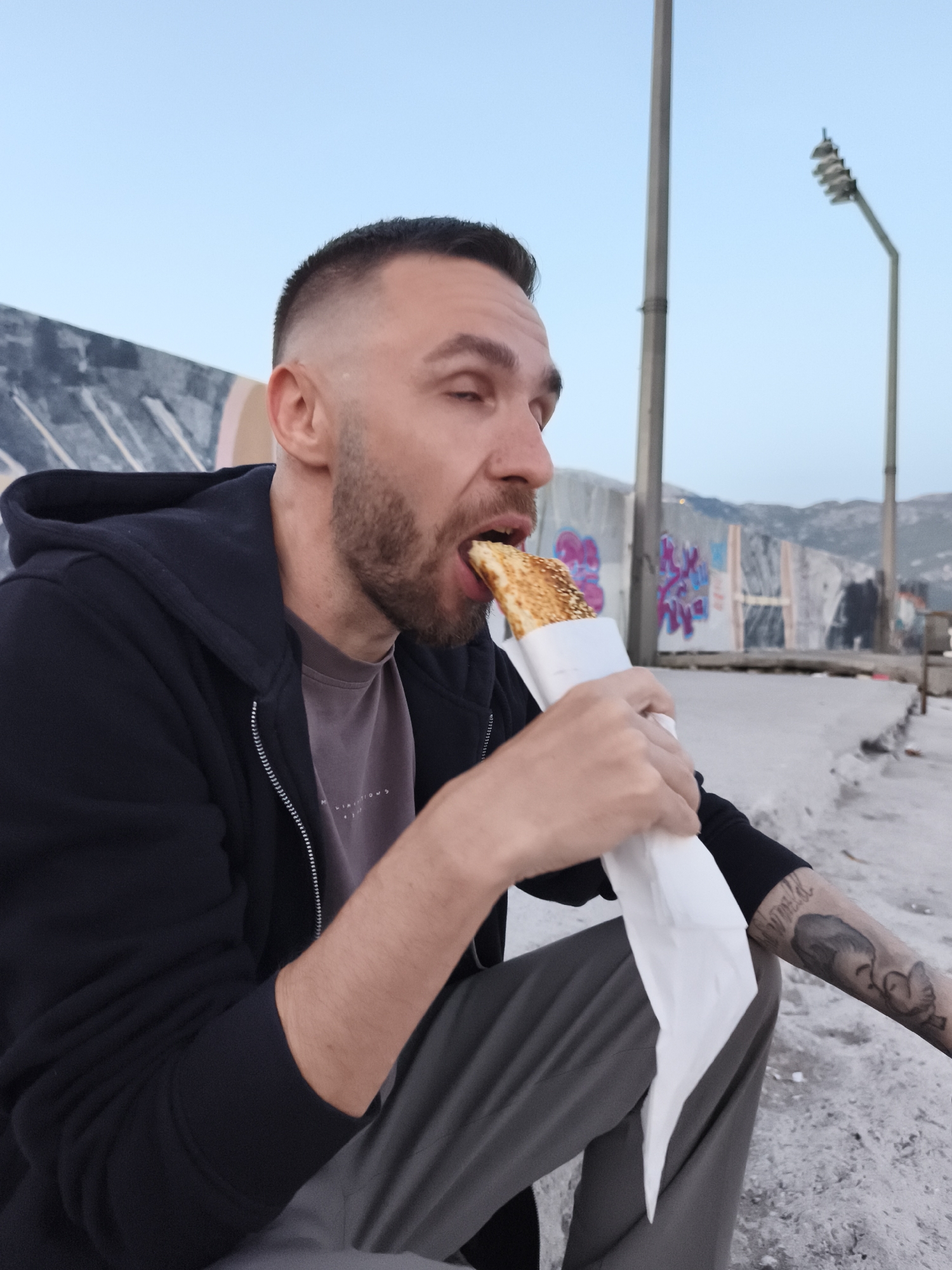 This outdoor portrait features a man in his late 20s or early 30s, with a short, faded haircut and a beard, actively eating. He is captured mid-bite, consuming a long, golden-brown pastry, possibly a burek or similar item, which is wrapped in white paper. His eyes are slightly closed in concentration as he takes the bite.
The man is dressed casually in a dark, presumably black, zip-up hooded jacket over a light grey or mauve t-shirt. Faint text, appearing to say "MLIMITATIONS," is visible on his t-shirt. He wears grey trousers. A tattoo, seemingly depicting a stylized face or figure with some accompanying script, is visible on his left forearm.
The setting appears to be an urban or semi-urban outdoor area in Budva, Montenegro, likely near a waterfront or industrial zone, given the concrete structures. The man is seated on a textured concrete surface. In the background, there is a tall concrete wall or barrier adorned with vibrant, multicolored graffiti, which includes abstract shapes and some illegible text. Beyond this wall, distant hills or mountains rise under a clear, pale blue sky. Several tall utility poles or streetlights punctuate the background, one with a distinct multi-tiered light fixture. The soft, even lighting suggests the time of day is either early morning or, more likely, late afternoon or early evening twilight. The weather is clear and calm. The scene conveys a moment of casual sustenance in a rugged, open environment.